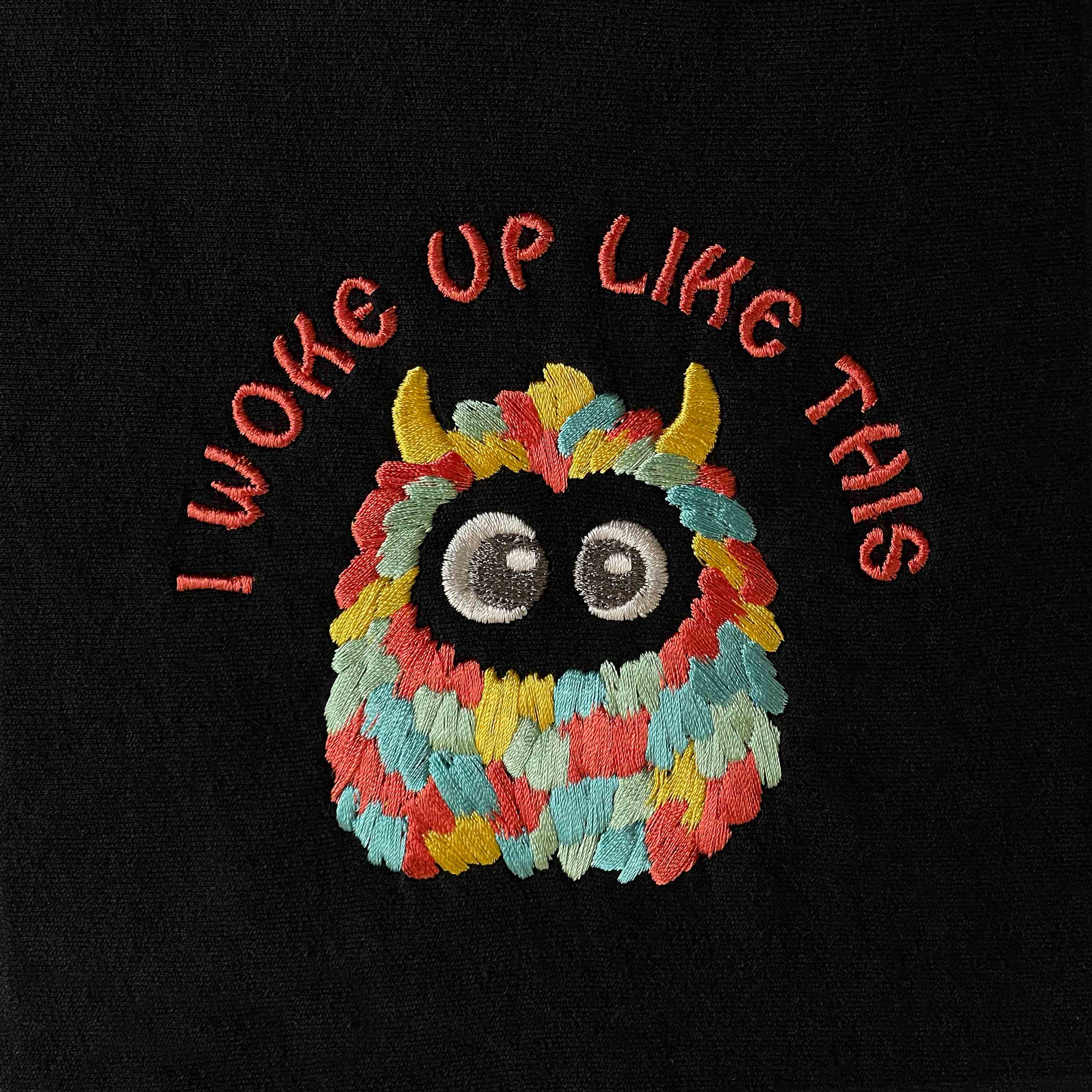 Siyah dokulu pamuklu kumaş üzerine işlenmiş "I WOKE UP LIKE THIS" nakışının makro (yakın çekim) detayı. Görselde sevimli, çok renkli ve tüylü canavar figürünün büyük gözlerinin ve kiremit tonlarındaki kavisli yazıların yoğun, sıkı dikiş dokusu net bir şekilde görülmektedir. İpliklerin canlılığı ve birinci sınıf Alman nakış işçiliğinin kalitesini gözler önüne seren, eğlenceli ve premium tasarımın yüksek çözünürlüklü detay karesi.