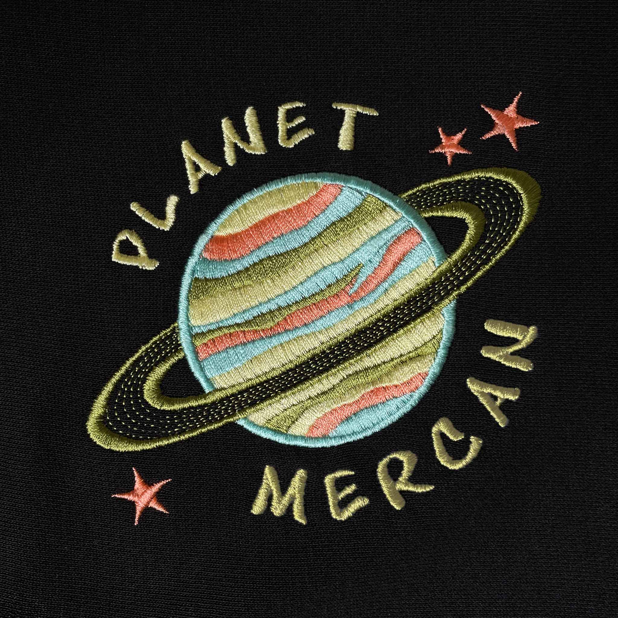 Siyah renkli isme özel çocuk kapüşonlu sweatshirt modelindeki gezegen tasarımının yakından görünümü. 'PLANET MERCAN' örneğiyle gösterilen yüksek kaliteli Alman nakış işçiliği, renkli ipliklerin kusursuz dokusunu gözler önüne serer. Oeko-Tex belgeli %100 pamuklu ağır gramajlı tok kumaş üzerine işlenen bu detaylar, yıkamaya karşı son derece dayanıklıdır ve çocukların hassas cildine zarar vermeyen sağlıklı bir yapıdadır.