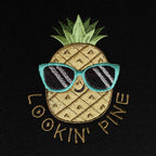 Siyah dokulu pamuklu kumaş üzerine işlenmiş "LOOKIN' PINE" ananas nakışının makro (yakın çekim) detayı. Görselde mavi çerçeveli güneş gözlüğü takan, gülümseyen ananas figürünün ve etrafındaki kiremit/gold tonlarındaki yazıların yoğun, sıkı dikiş dokusu net bir şekilde görülmektedir. İpliklerin canlılığı ve birinci sınıf Alman nakış işçiliğinin kalitesini gözler önüne seren, eğlenceli ve premium tasarımın yüksek çözünürlüklü detay karesi.