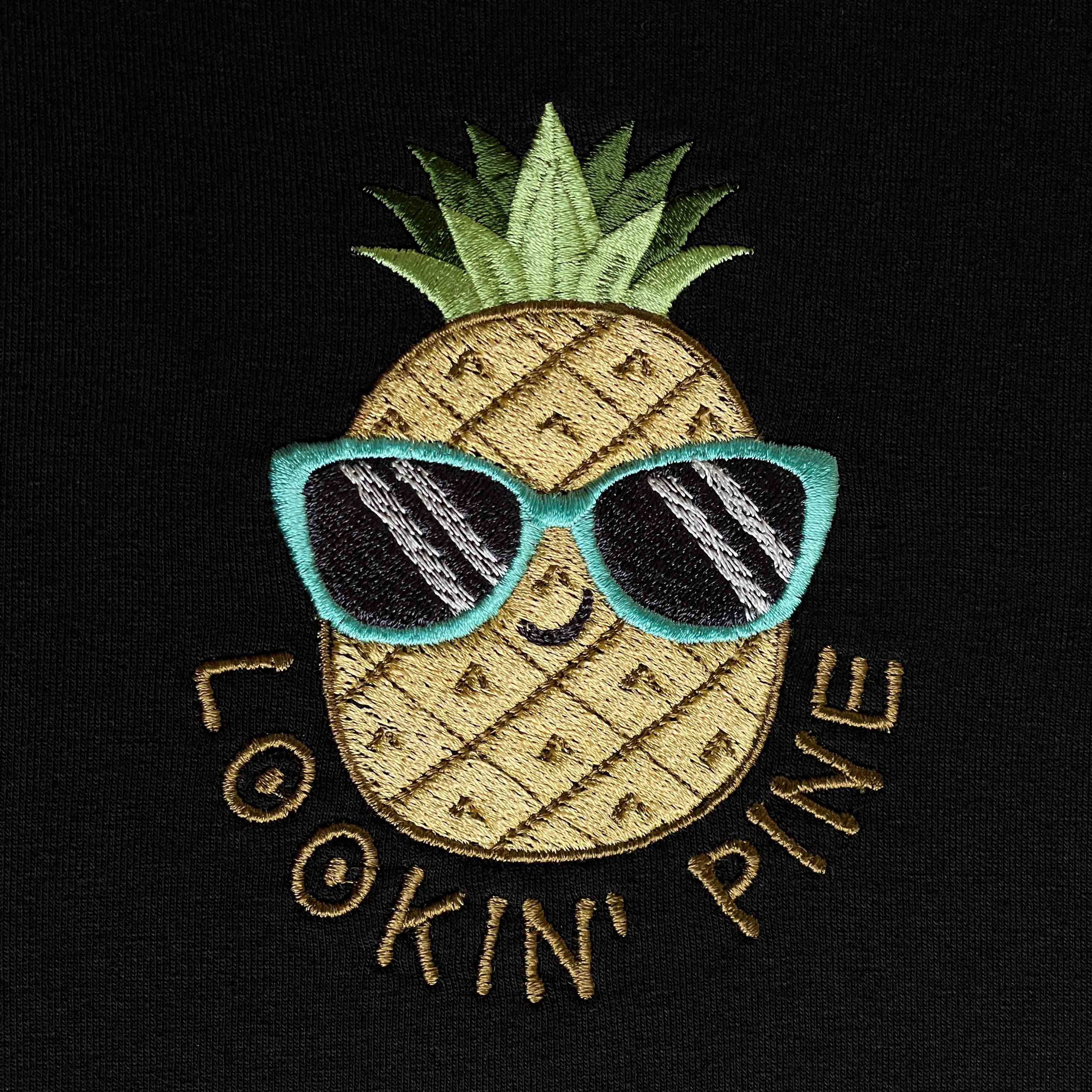 Siyah dokulu pamuklu kumaş üzerine işlenmiş "LOOKIN' PINE" ananas nakışının makro (yakın çekim) detayı. Görselde mavi çerçeveli güneş gözlüğü takan, gülümseyen ananas figürünün ve etrafındaki kiremit/gold tonlarındaki yazıların yoğun, sıkı dikiş dokusu net bir şekilde görülmektedir. İpliklerin canlılığı ve birinci sınıf Alman nakış işçiliğinin kalitesini gözler önüne seren, eğlenceli ve premium tasarımın yüksek çözünürlüklü detay karesi.