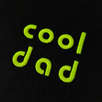 Siyah dokulu pamuklu kumaş üzerine, floresan özellikli neon sarı (neon sarı) iplikle işlenmiş "cool dad" nakışının makro (yakın çekim) detayı. Görselde nakışın blok ve yuvarlak yazı tipindeki harflerinin yoğun, sıkı dikiş dokusu, ipliklerin canlılığı ve birinci sınıf Alman nakış ipliği net bir şekilde görülmektedir. Premium babalar için tasarlanmış giyim parçalarının kalitesini vurgulayan yüksek çözünürlüklü detay karesi.