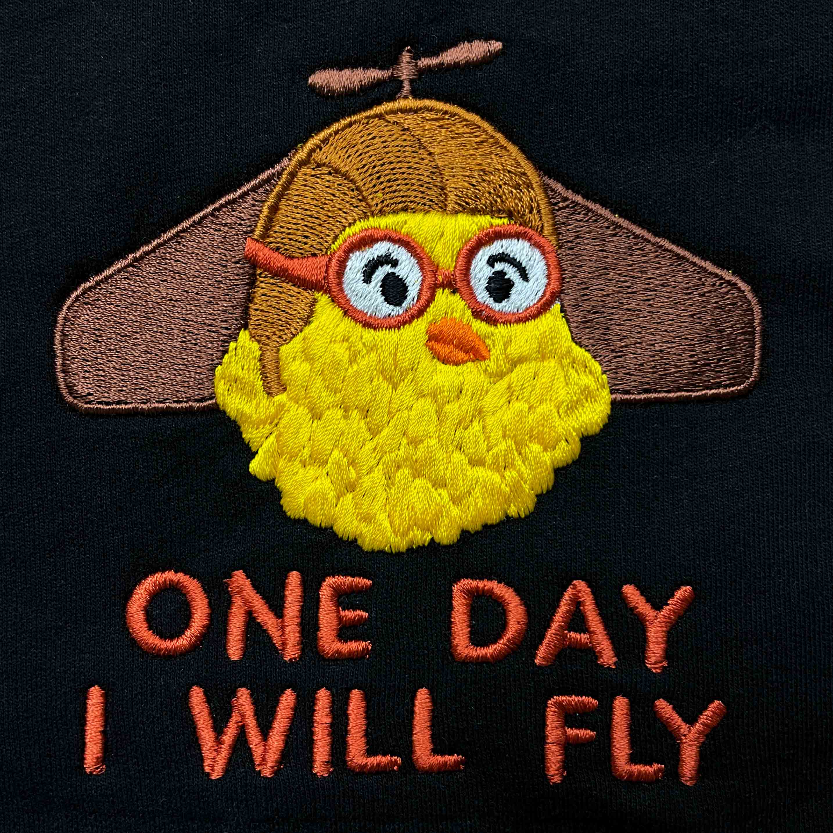 Siyah dokulu pamuklu kumaş üzerine işlenmiş "ONE DAY I WILL FLY" nakışının makro (yakın çekim) detayı. Görselde kahverengi havacı şapkası ve kırmızı çerçeveli pilot gözlüğü takan sevimli sarı civciv figürünün, şapka üzerindeki pervanenin ve kiremit tonlarındaki yazıların yoğun, sıkı dikiş dokusu net bir şekilde görülmektedir. İpliklerin canlılığı ve birinci sınıf Alman nakış işçiliğinin kalitesini gözler önüne seren, eğlenceli ve premium tasarımın yüksek çözünürlüklü detay karesi.