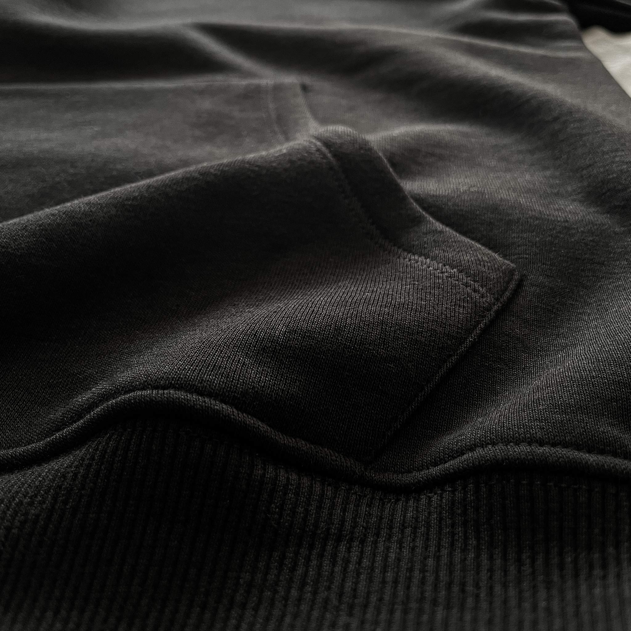 Siyah premium yetişkin pullover hoodie (kapüşonlu sweatshirt) modelinin kanguru cebinin bel ribanasıyla (alt bant) birleştiği noktayı gösteren makro detay çekimi. Kaliteli tok pamuklu kumaşın yoğun örgüsü ile esnek fitilli ribananın sağlam dikişlerle buluştuğu kusursuz işçilik. Cebin alt kısmındaki dayanıklılığı ve temiz bitişi vurgulayan premium yapısal detay.