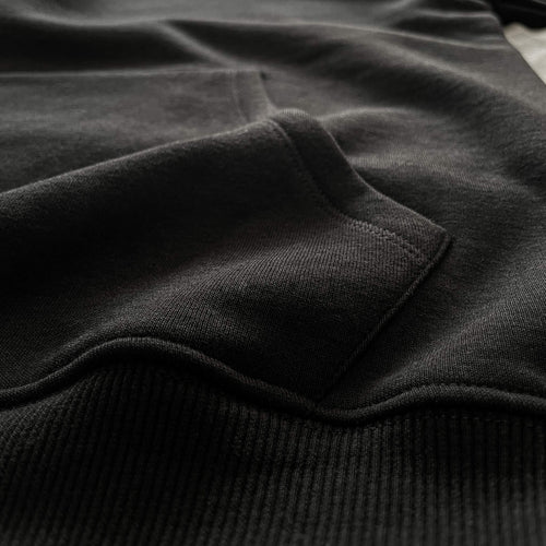 Siyah premium yetişkin pullover hoodie (kapüşonlu sweatshirt) modelinin kanguru cebinin bel ribanasıyla (alt bant) birleştiği noktayı gösteren makro detay çekimi. Kaliteli tok pamuklu kumaşın yoğun örgüsü ile esnek fitilli ribananın sağlam dikişlerle buluştuğu kusursuz işçilik. Cebin alt kısmındaki dayanıklılığı ve temiz bitişi vurgulayan premium yapısal detay.