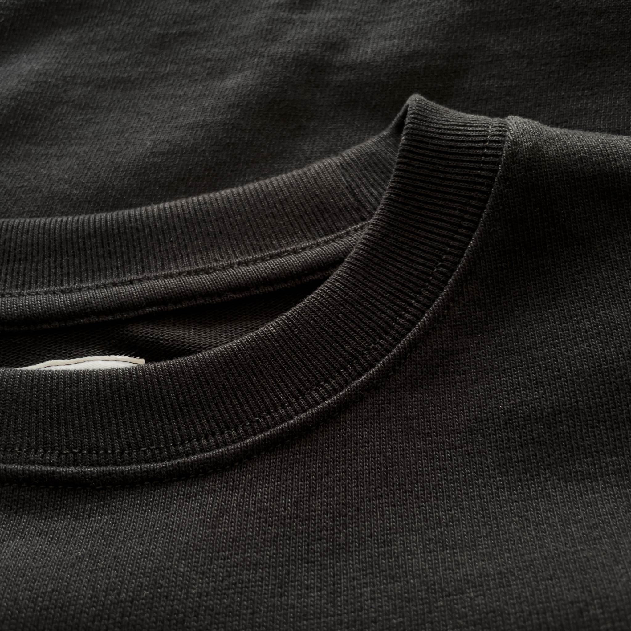Siyah premium yetişkin t-shirt modelinin esnek fitilli yaka ribanasını ve tok pamuklu kumaş dokusunu gösteren yakın çekim. Kaliteli ağır gramajlı (heavyweight) kumaşın yoğun örgüsü boynu nazikçe sarar ve tok duruşuyla formunu korur. Minimalist şıklık ve premium konfor sunan yapısal detay.