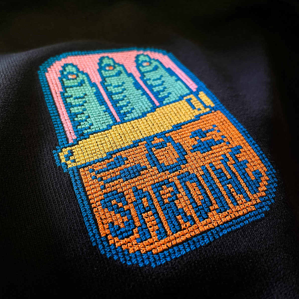 Siyah pamuklu tişört kumaşı üzerine işlenmiş "Sardine" (sardalya konservesi) nakışının makro (yakın çekim) detayı. Retro pixel art estetiği ve geleneksel kanaviçe (cross stitch) dokusunun birleştiği tasarımda; siyah zemin üzerinde patlayan pembe, turkuaz, sarı ve turuncu renkli ipliklerin kusursuz dizilimi vurgulanmaktadır. Birinci sınıf Alman nakış ipliği kullanılarak özenle oluşturulmuş, nostaljik ve premium tasarımın yüksek çözünürlüklü detay karesi.