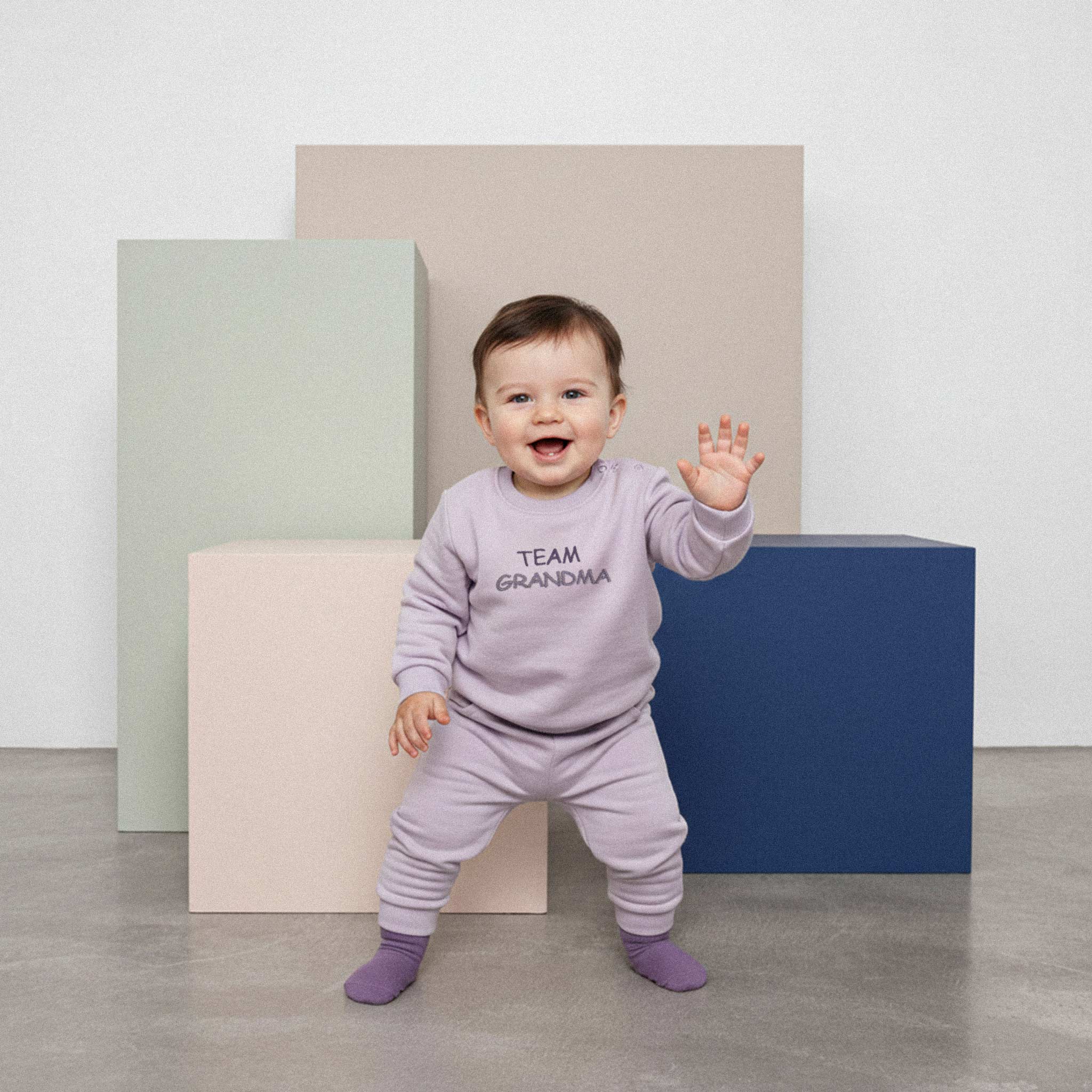 Üzerinde lila renkli, 'Team Grandma' nakışlı eşofman takımı bulunan sevimli bebek, renkli küplerin önünde durarak neşeyle el sallıyor.
