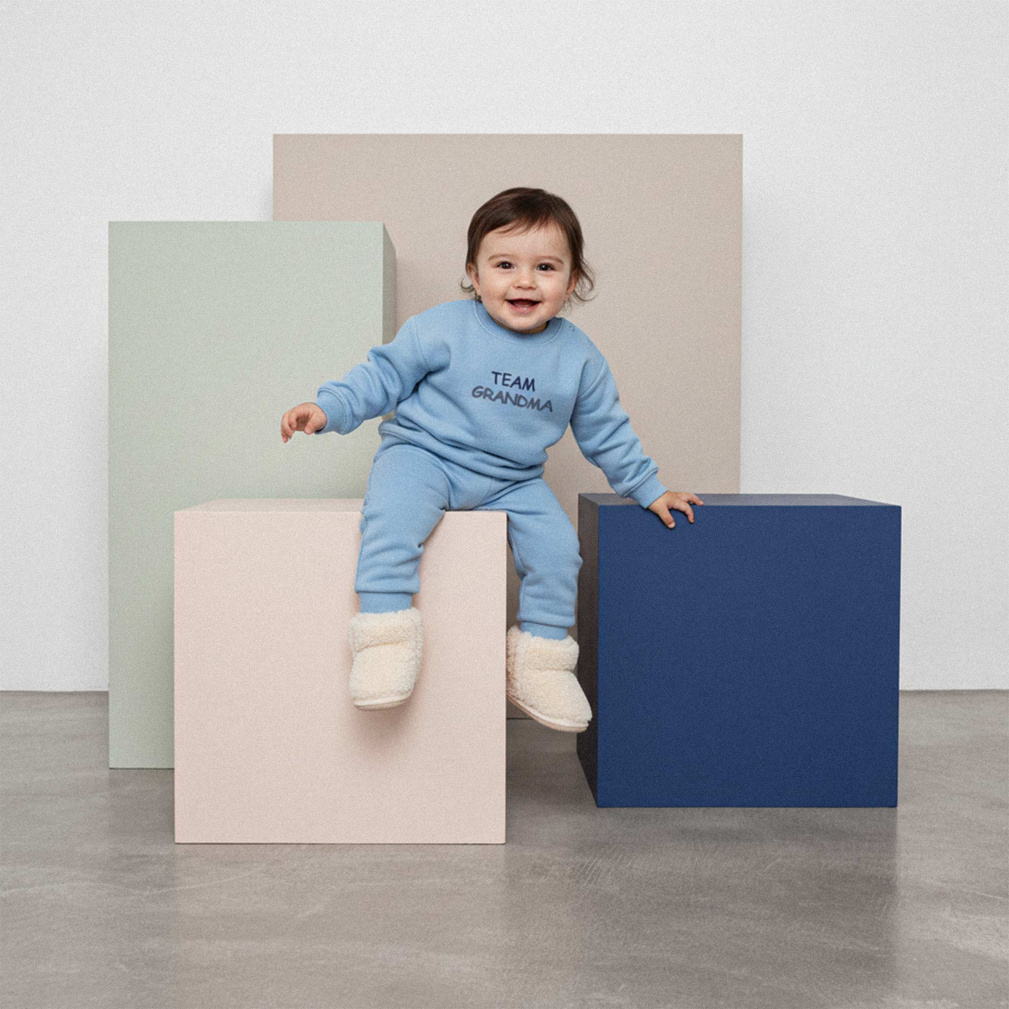 Üzerinde mavi renkli, 'Team Grandma' nakışlı eşofman takımı ve tüylü patikler bulunan sevimli bebek, renkli bir küpün üzerinde otururken neşeyle gülümsüyor.