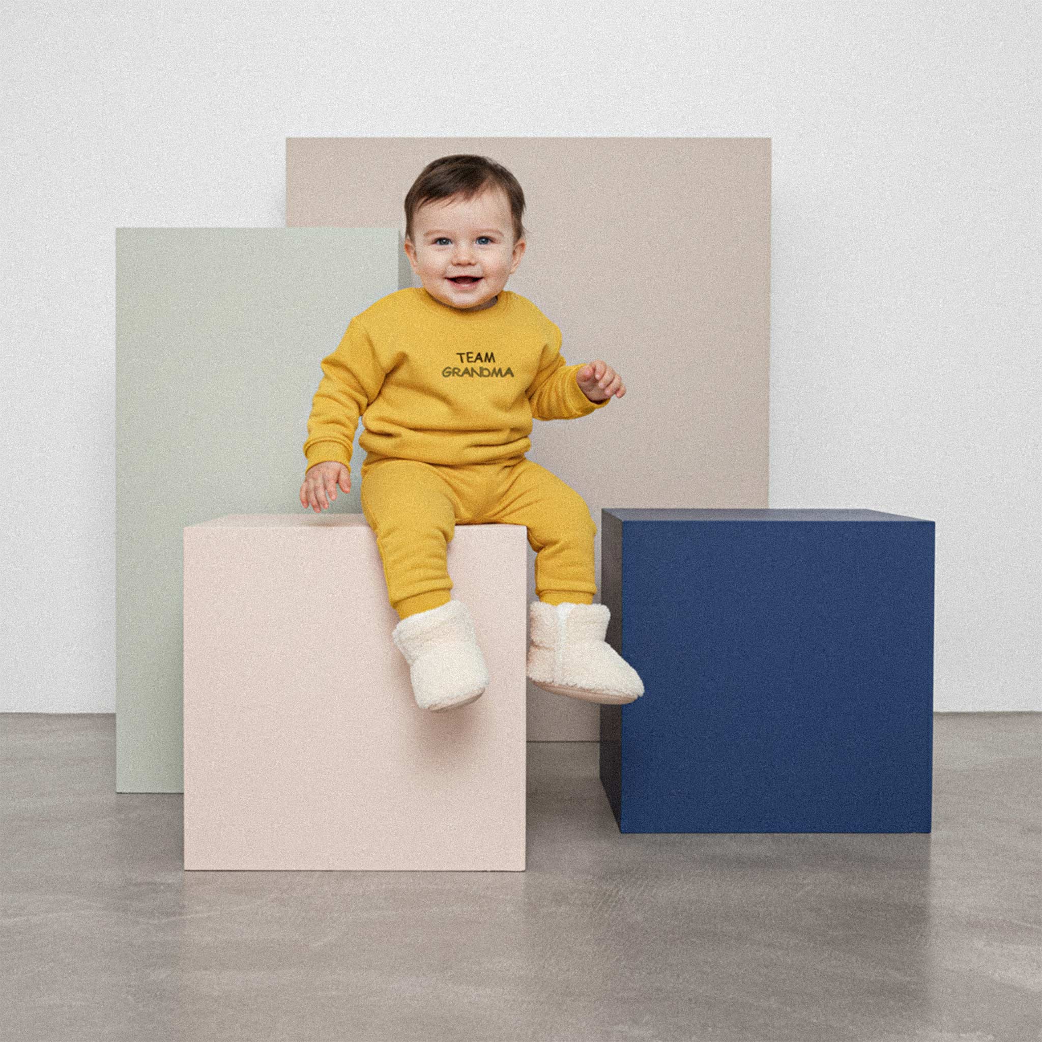 Üzerinde hardal sarısı renkli, 'Team Grandma' nakışlı eşofman takımı ve tüylü patikler bulunan sevimli bebek, renkli küplerin üzerinde otururken neşeyle gülümsüyor.