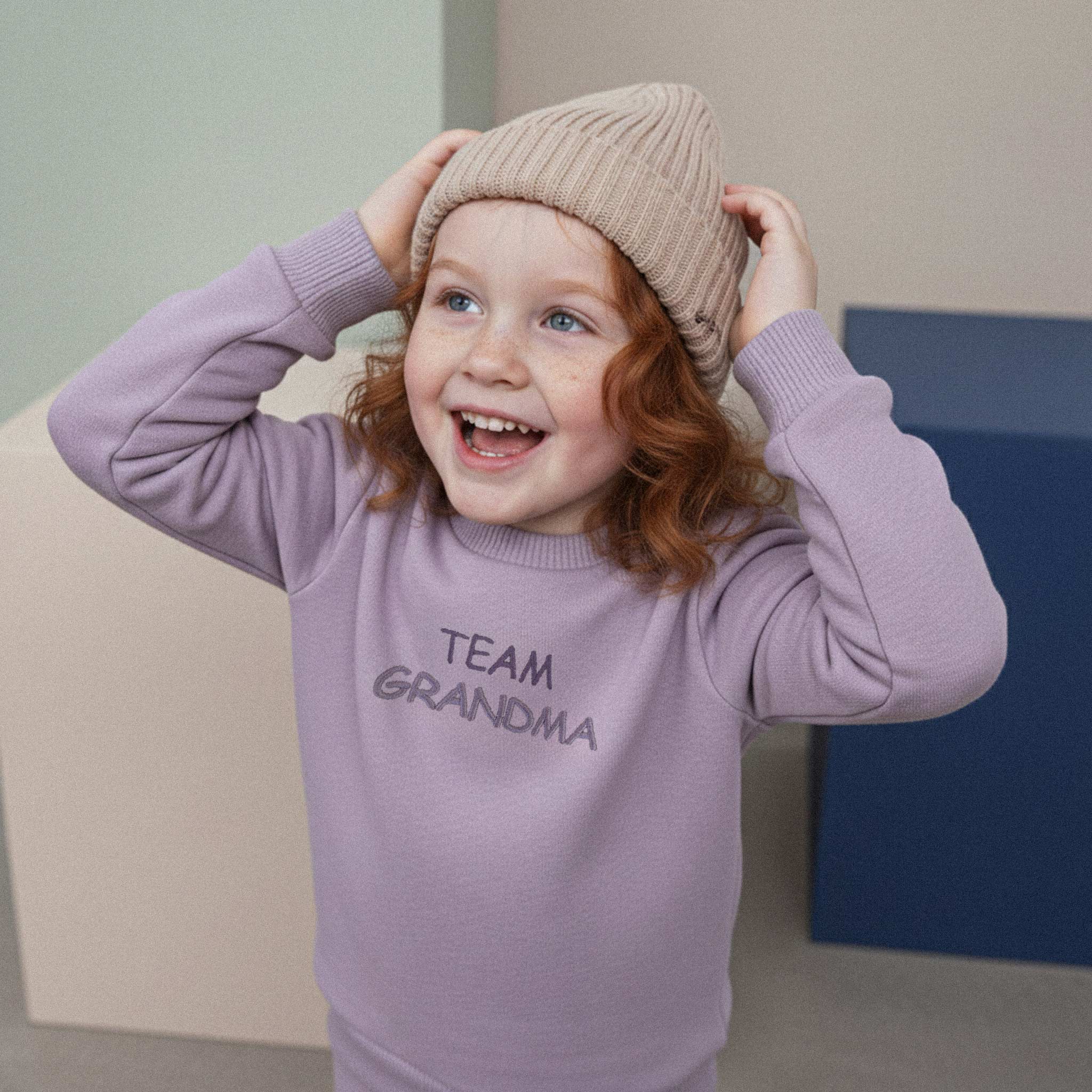 Kızıl saçlı, lila renkli 'Team Grandma' eşofman takımı ve bej bere giyen kız çocuk, beresini tutarken neşeyle kahkaha atıyor.