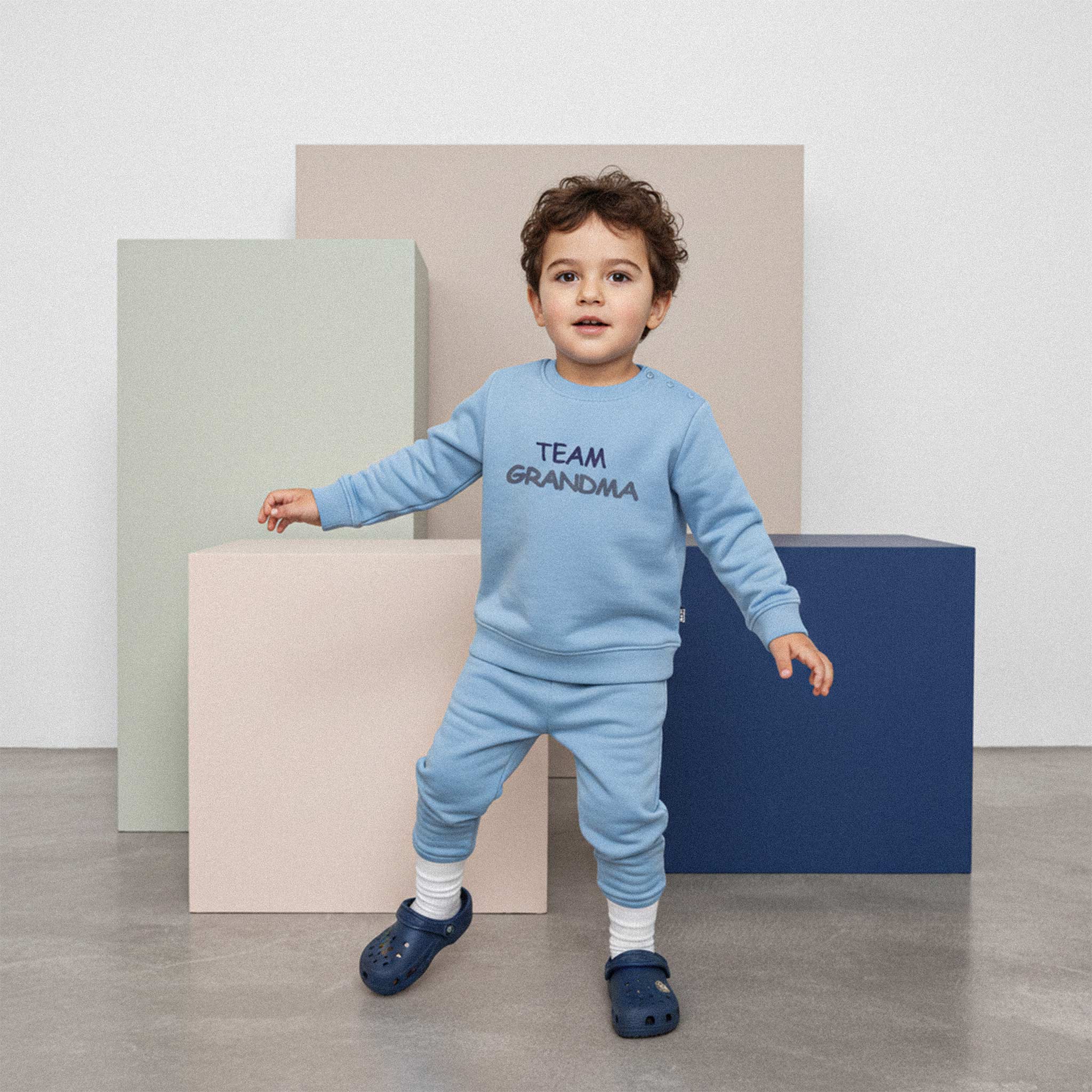 Kıvırcık saçlı, mavi renkli 'Team Grandma' eşofman takımı giyen erkek çocuk, renkli küplerin önünde durarak enerjik bir şekilde poz veriyor.