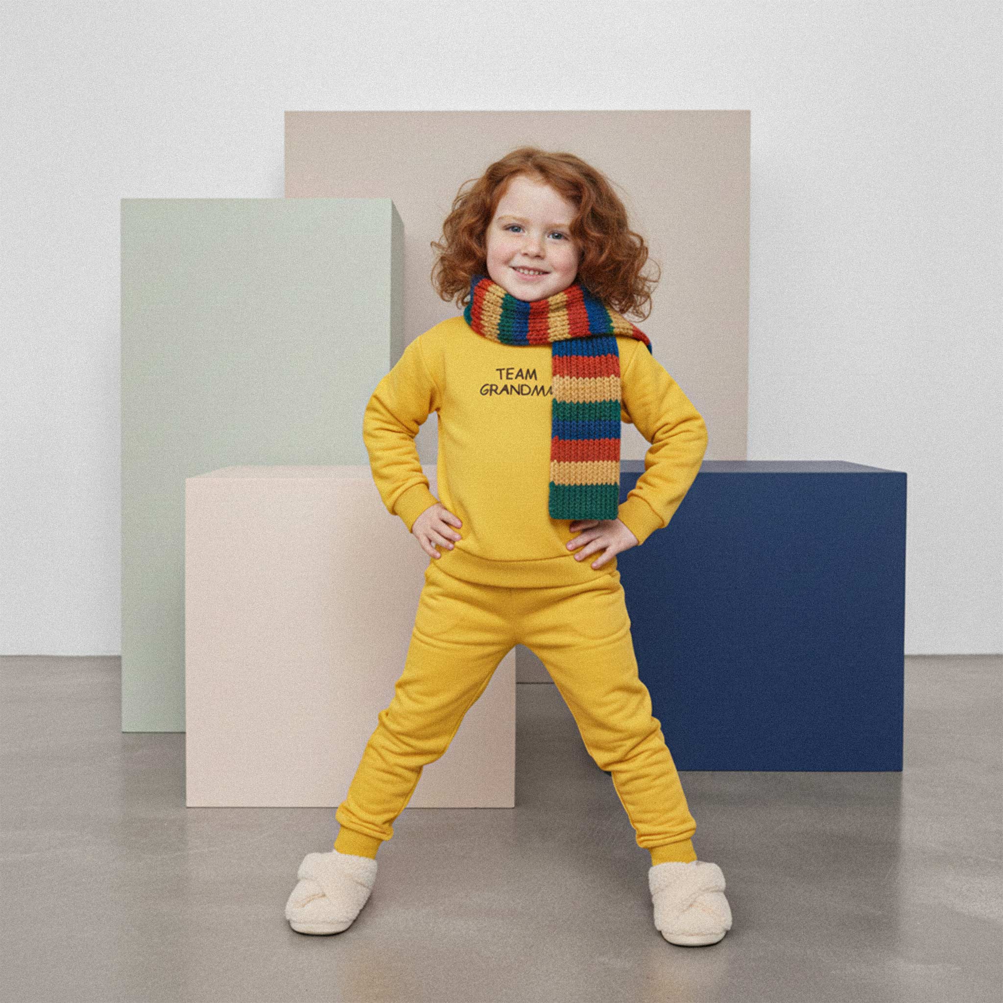 Kızıl saçlı, hardal sarısı 'Team Grandma' eşofman takımı ve renkli bir atkı takan kız çocuk, elleri belinde poz vererek gülümsüyor.