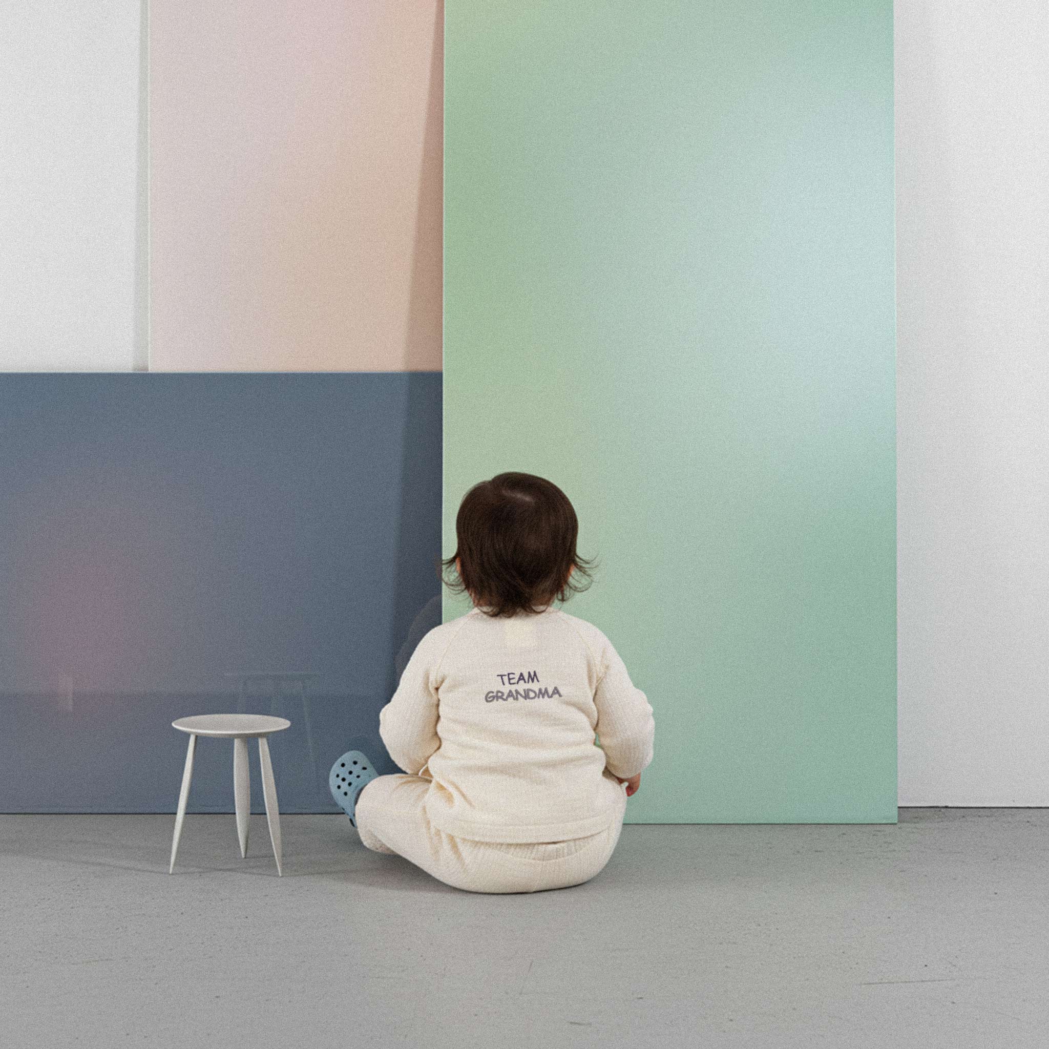 Üzerinde ekru renkli, 'Team Grandma' nakışlı müslin kimono takımı bulunan sevimli bebek, renkli küplerin önünde yerde otururken neşeyle gülümsüyor.