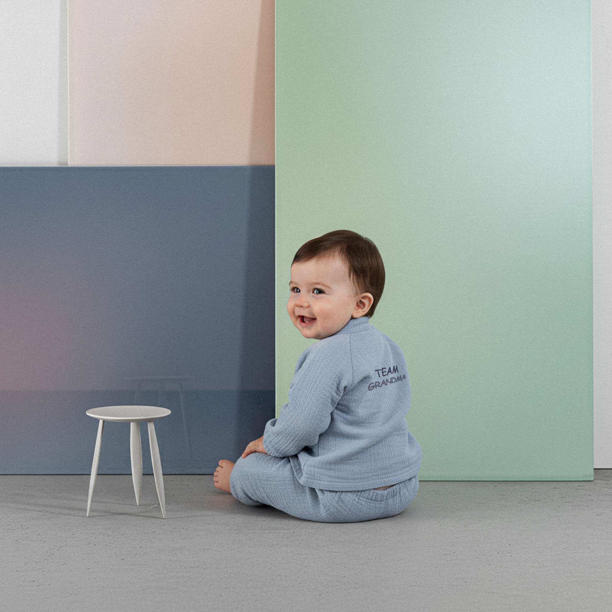 Üzerinde indigo mavisi renkli, 'Team Grandma' nakışlı müslin kimono takımı bulunan sevimli bebek, renkli küplerin önünde yerde otururken kollarını açarak neşeyle gülümsüyor.