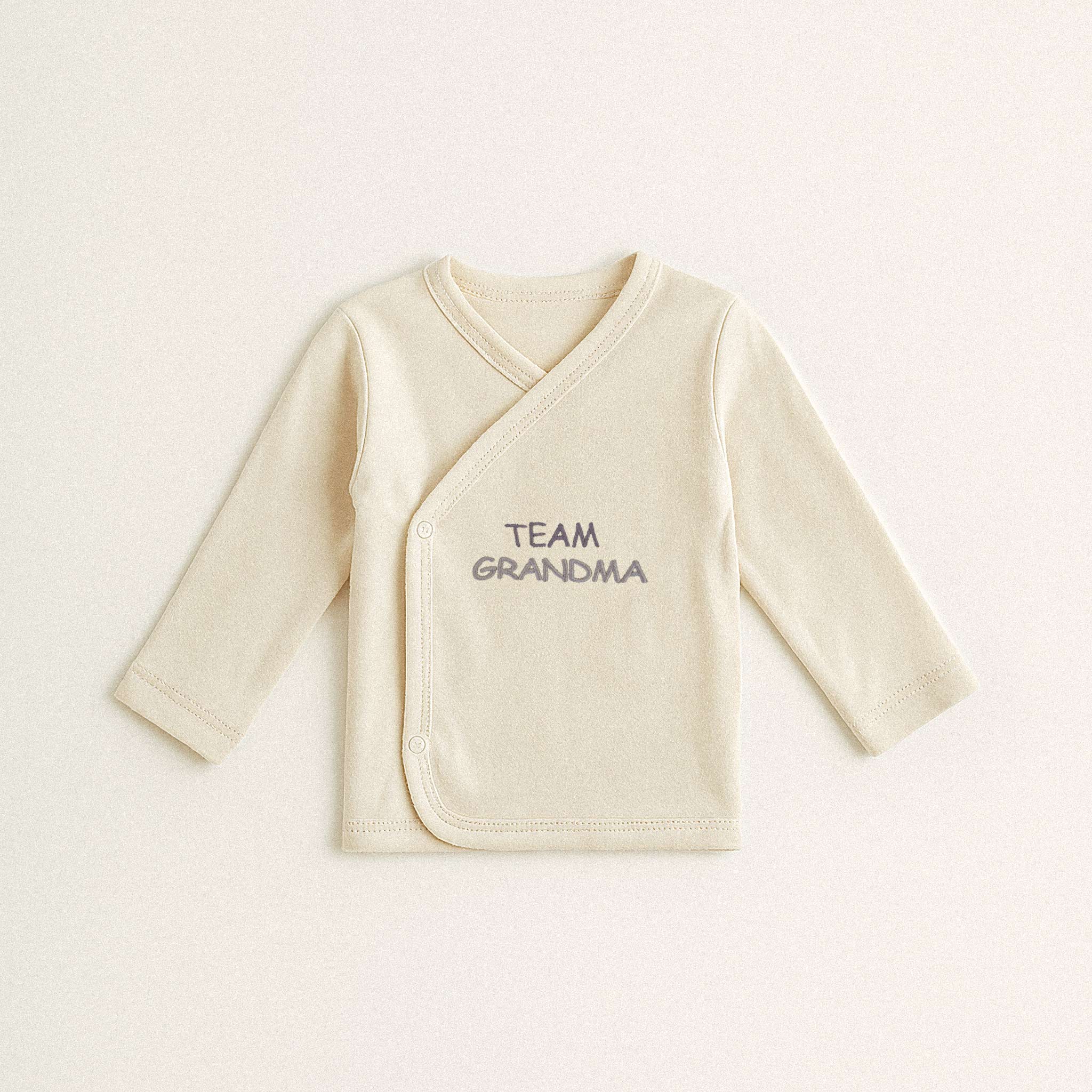 Beyaz arka plan üzerinde sergilenen, ekru renkli, %100 pamuklu, uzun kollu kruvaze yenidoğan üst. Üzerinde lila rengi iplikle 'TEAM GRANDMA' yazısı işlenmiştir.