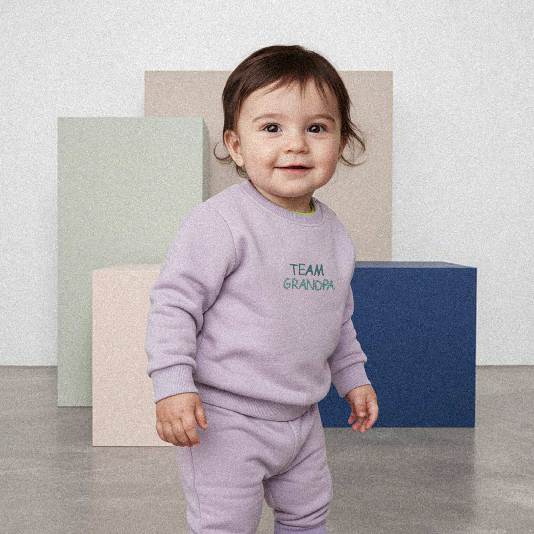 Üzerinde lila renkli, 'Team Grandpa' nakışlı eşofman takımı bulunan sevimli bebek, renkli küplerin önünde durarak neşeyle gülümsüyor.