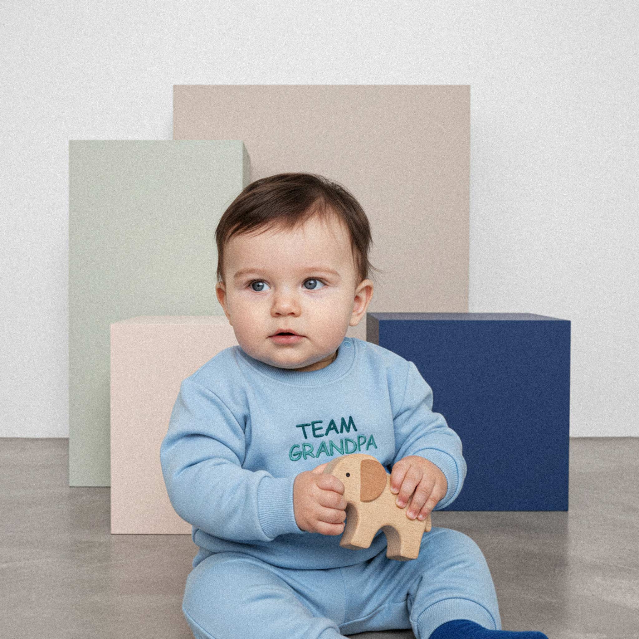 Üzerinde mavi renkli, 'Team Grandpa' nakışlı eşofman takımı bulunan sevimli bebek, elindeki ahşap fil oyuncağıyla yerde oturuyor.