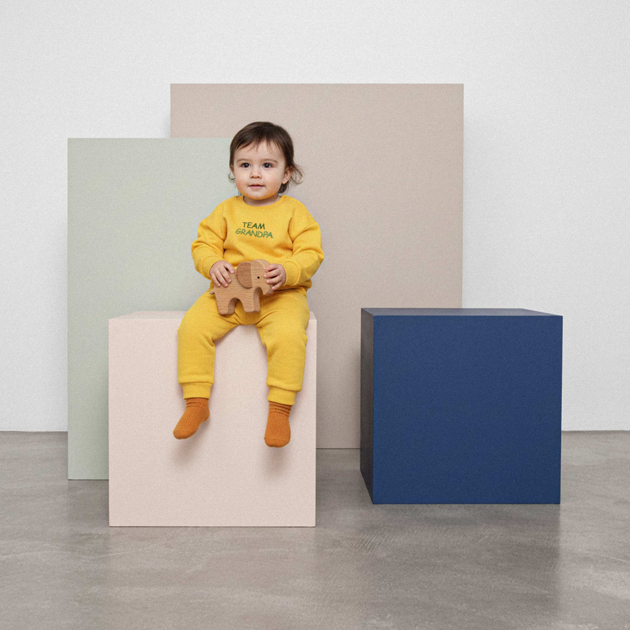 Üzerinde hardal sarısı renkli, 'Team Grandpa' nakışlı eşofman takımı bulunan sevimli bebek, elindeki ahşap fil oyuncağıyla renkli bir küpün üzerinde oturuyor.
