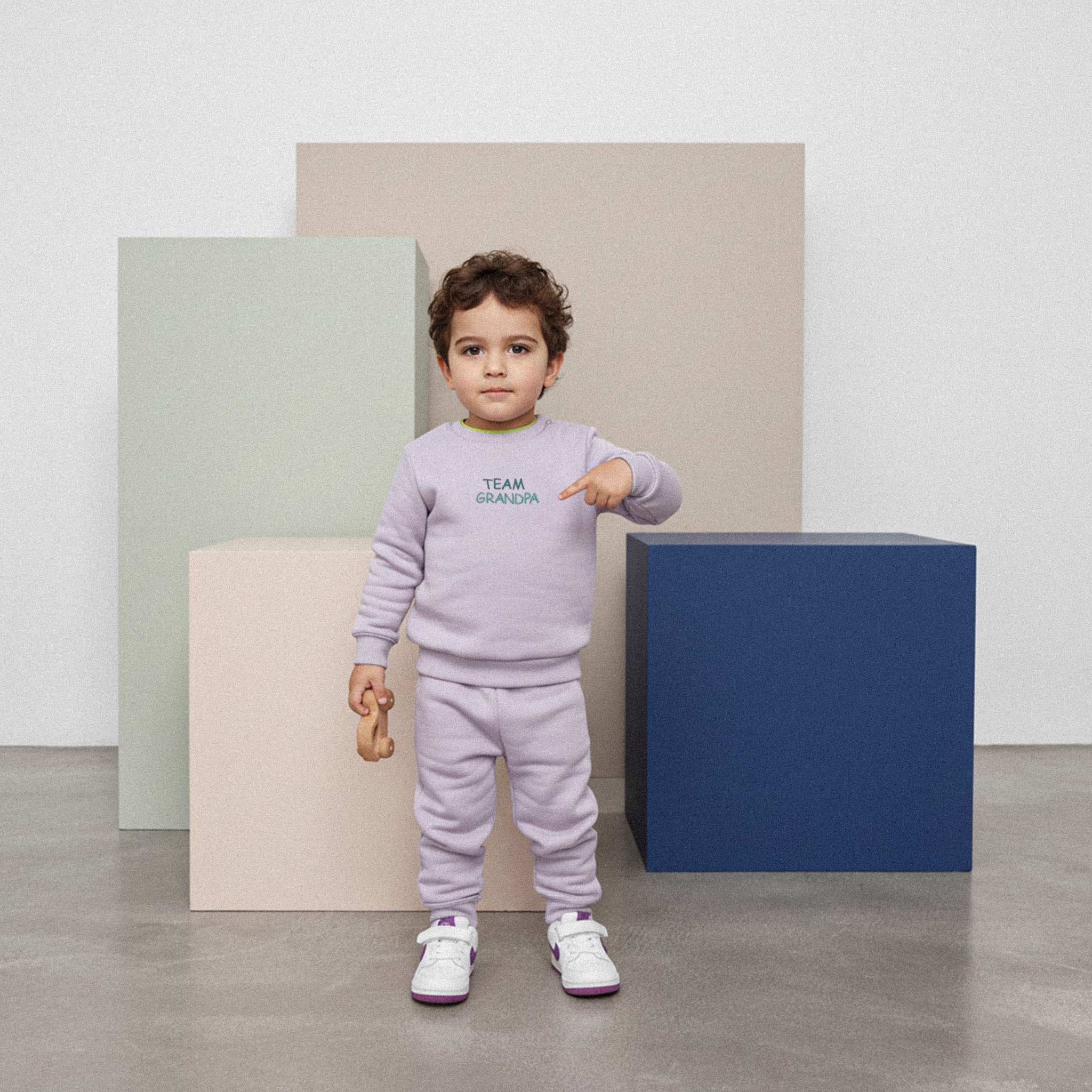 Kıvırcık saçlı, lila renkli 'Team Grandpa' eşofman takımı giyen erkek çocuk, elindeki oyuncakla dururken parmağıyla sweatshirt üzerindeki nakışı işaret ediyor.