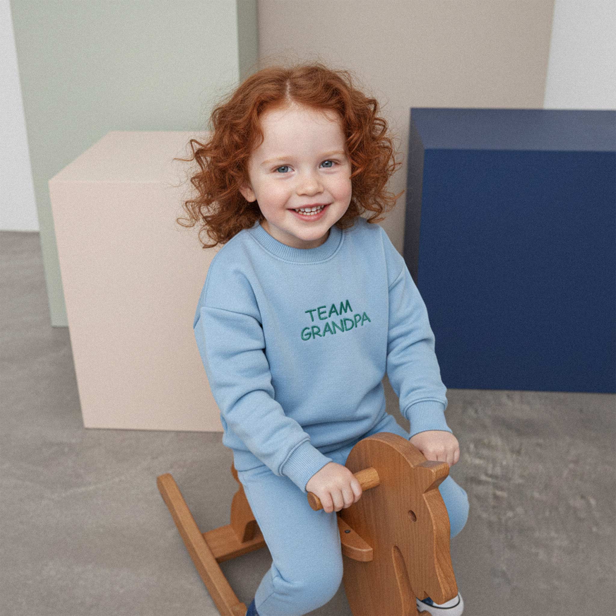 Kızıl saçlı, mavi renkli 'Team Grandpa' eşofman takımı giyen çocuk, ahşap bir sallanan at üzerinde otururken neşeyle gülümsüyor.