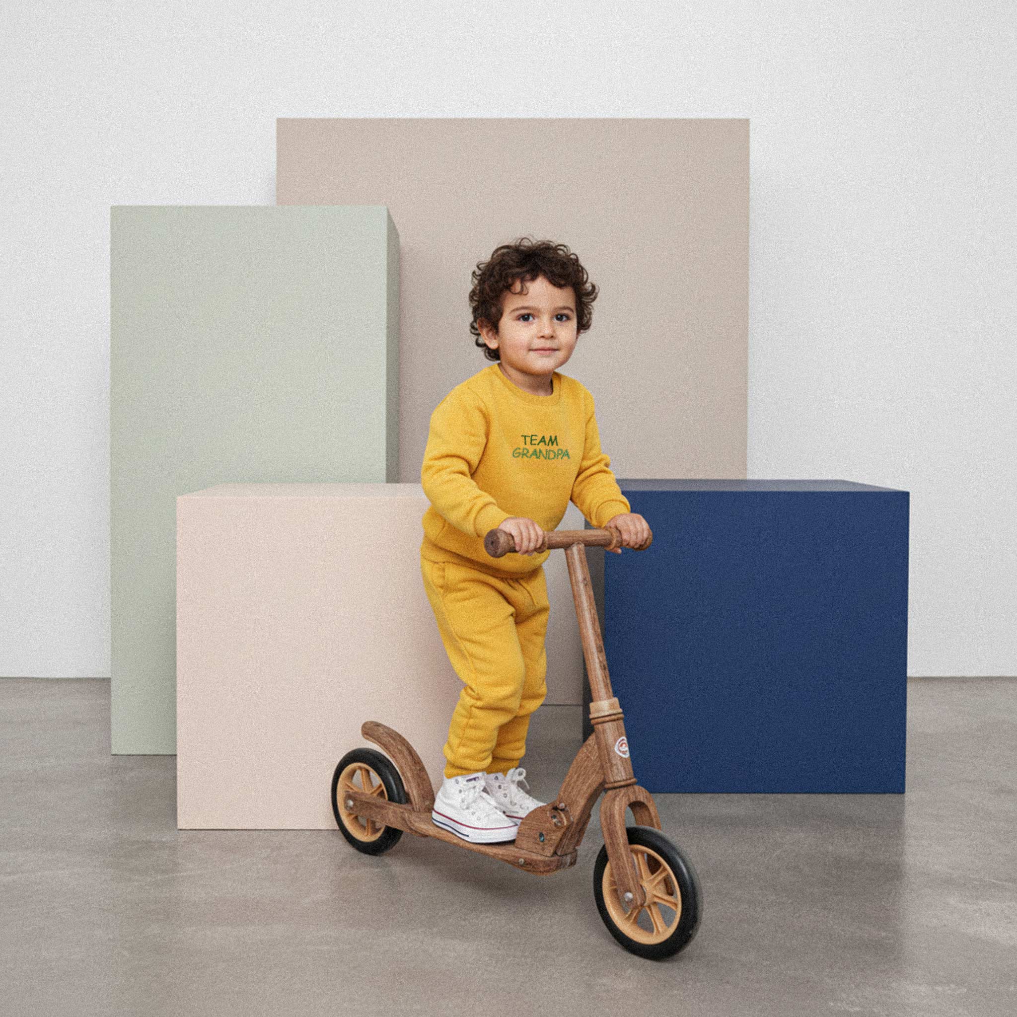 Kıvırcık saçlı, hardal sarısı 'Team Grandpa' eşofman takımı giyen erkek çocuk, ahşap bir scooter üzerinde durarak sevimli bir şekilde poz veriyor.