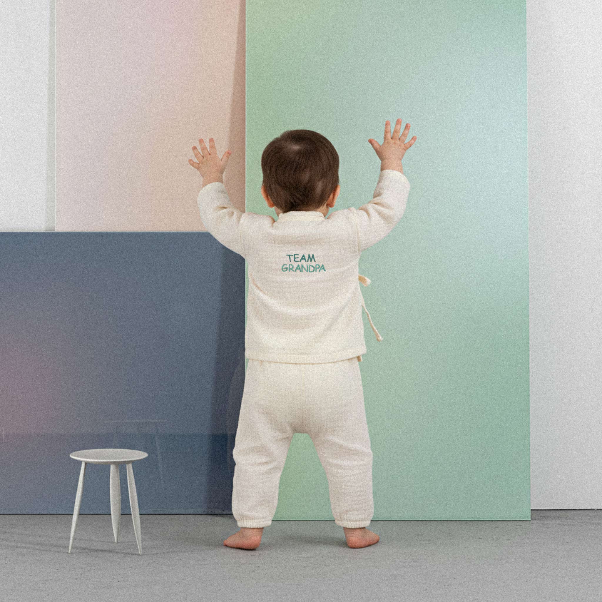 Üzerinde ekru renkli, 'Team Grandpa' nakışlı müslin kimono takımı bulunan sevimli bebek, renkli küplerin önünde ayakta durarak neşeyle ellerini kaldırıyor.
