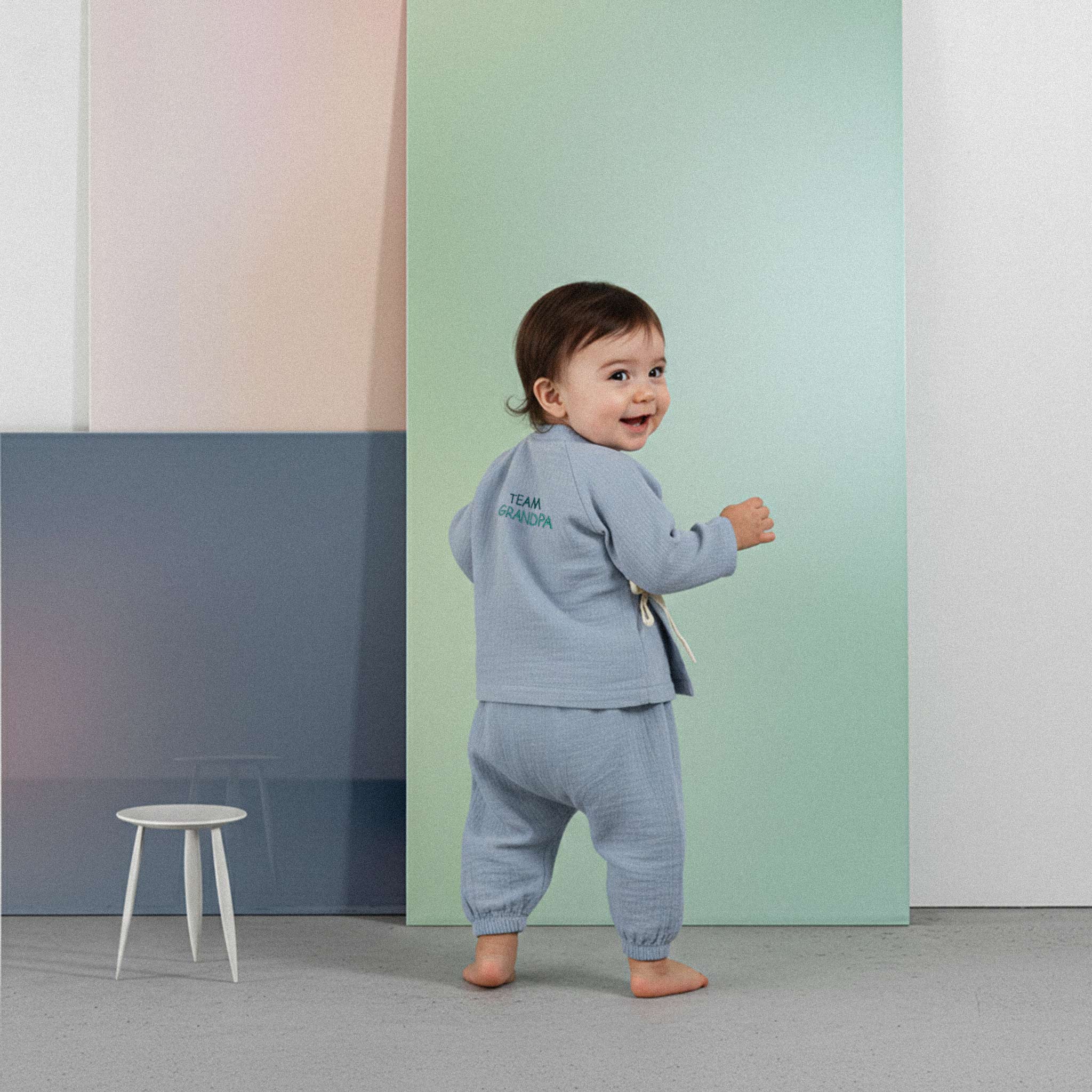 Üzerinde indigo mavisi renkli, 'Team Grandpa' nakışlı müslin kimono takımı bulunan sevimli bebek, renkli küplerin önünde yürümeye çalışırken neşeyle gülümsüyor.