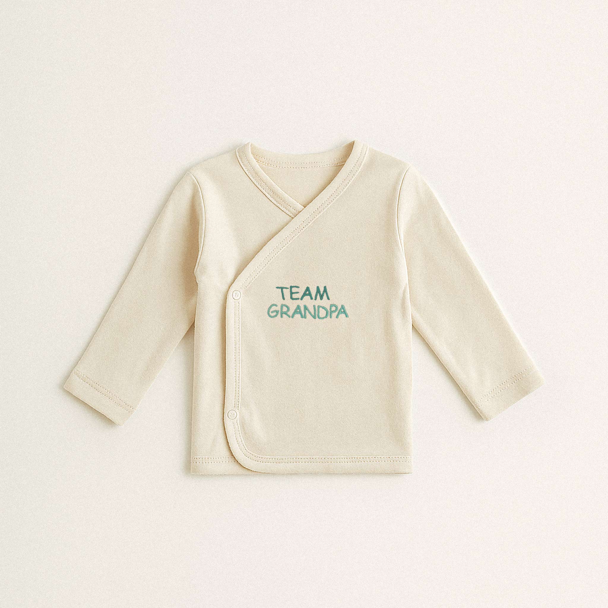 Beyaz arka plan üzerinde duran, ekru renkli, organik pamuklu, uzun kollu kruvaze yenidoğan üst. Üzerinde yeşil iplikle 'TEAM GRANDPA' yazısı işlenmiştir.