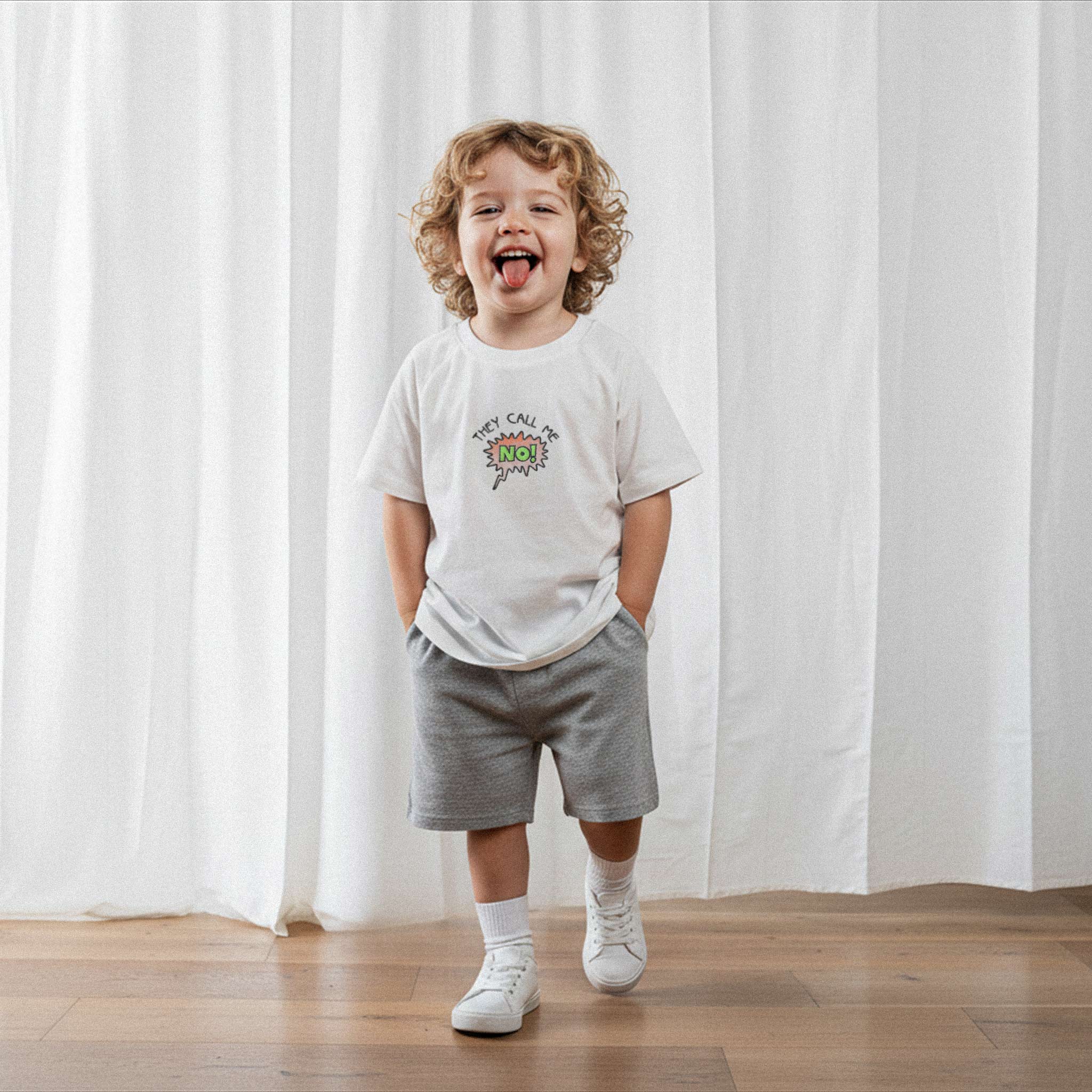 Gri şort ve ekru renkli, 'They Call Me No!' nakışlı reglan kollu oversize bebek tişörtü giyen kıvırcık saçlı küçük erkek çocuk, dilini çıkararak neşeyle poz veriyor.