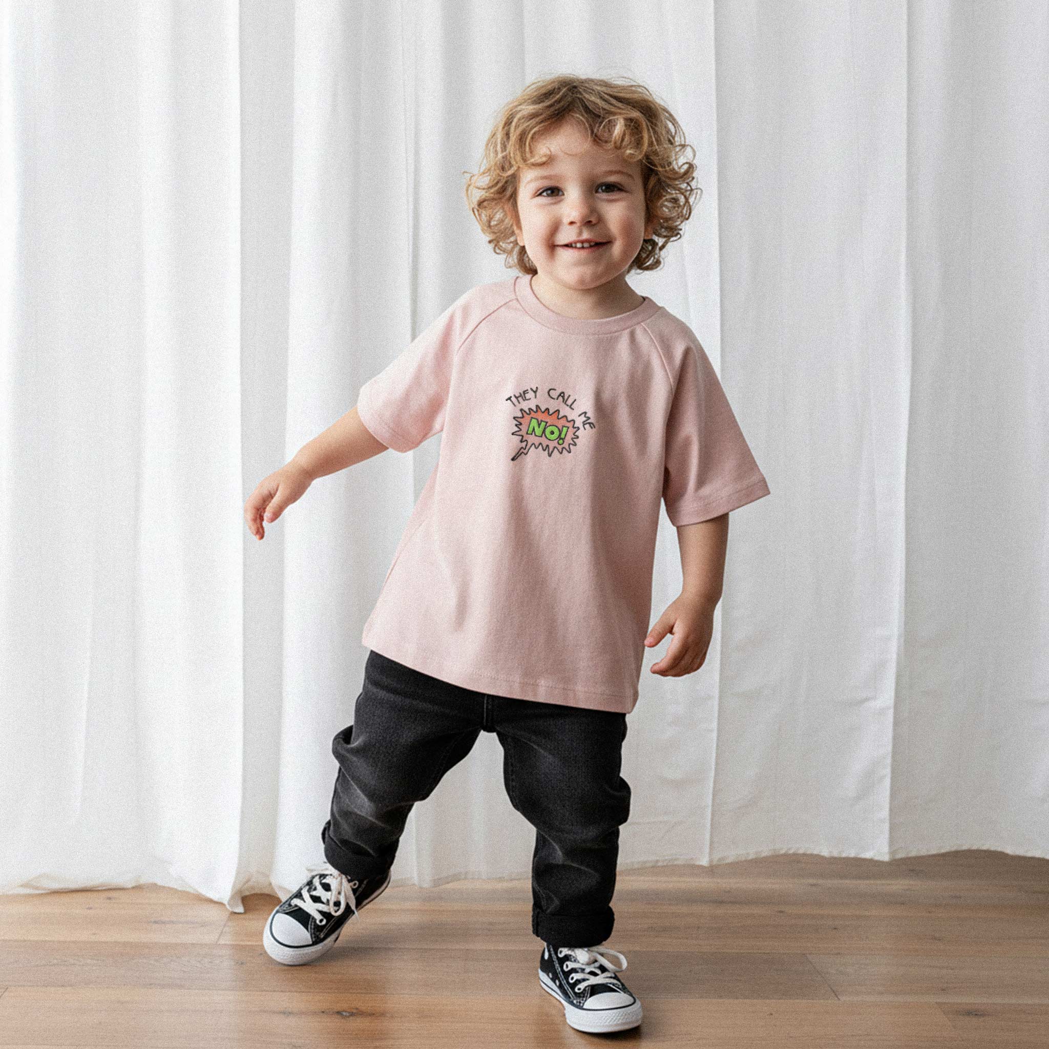Siyah pantolon ve gül kurusu renkli, 'They Call Me No!' nakışlı reglan kollu oversize bebek tişörtü giyen kıvırcık saçlı küçük erkek çocuk, sevimli bir şekilde gülümseyerek poz veriyor.