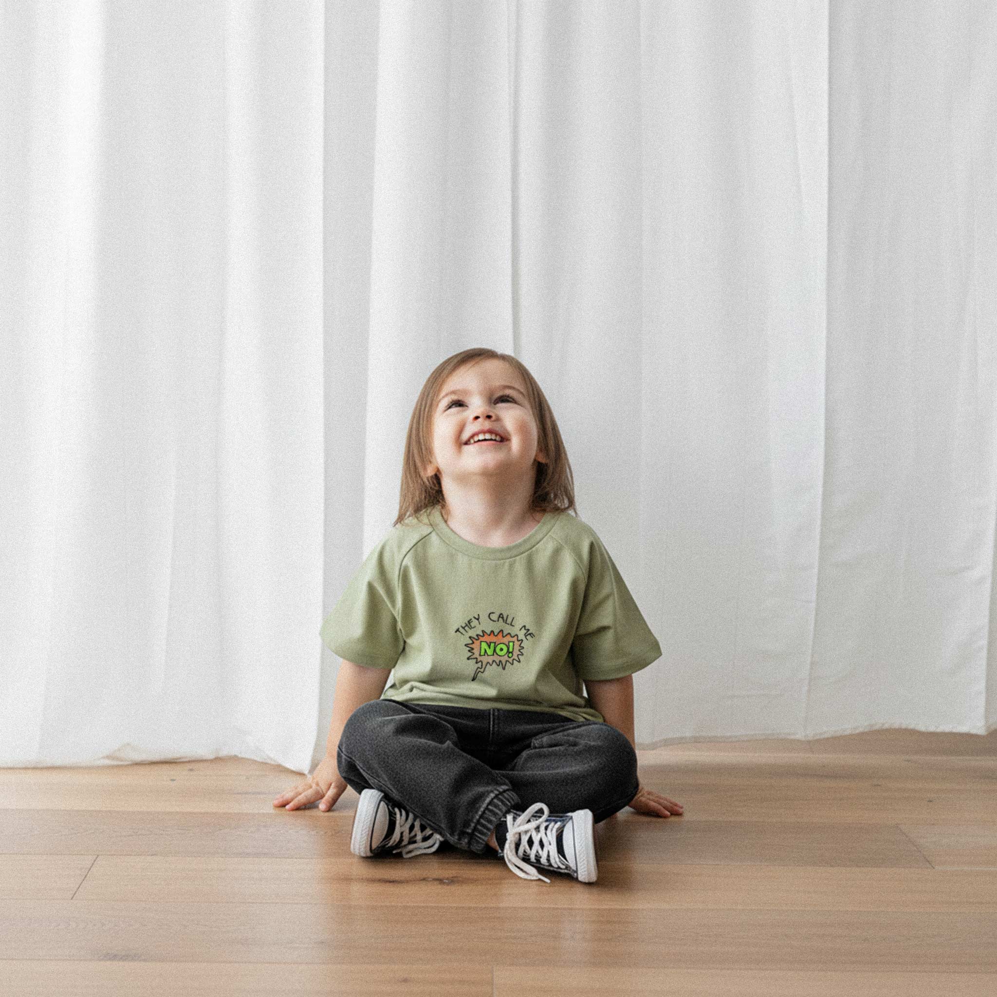 Siyah pantolon ve haki yeşili, 'They Call Me No!' nakışlı reglan kollu oversize bebek tişörtü giyen küçük kız çocuk, yerde otururken neşeyle yukarı bakıyor.