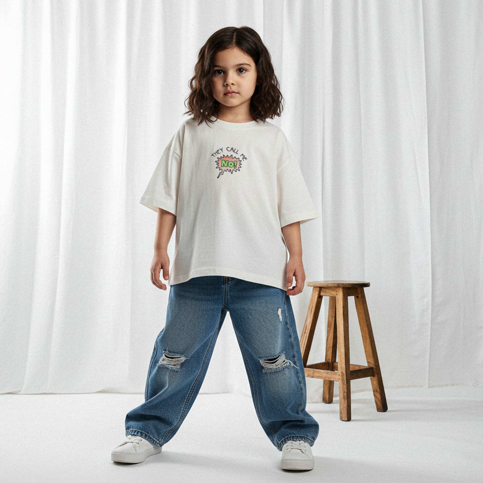 Yırtık jean pantolon ve ekru renkli, 'They Call Me No!' nakışlı oversize tişört giyen kız çocuk, kendinden emin bir duruşla kameraya bakıyor.