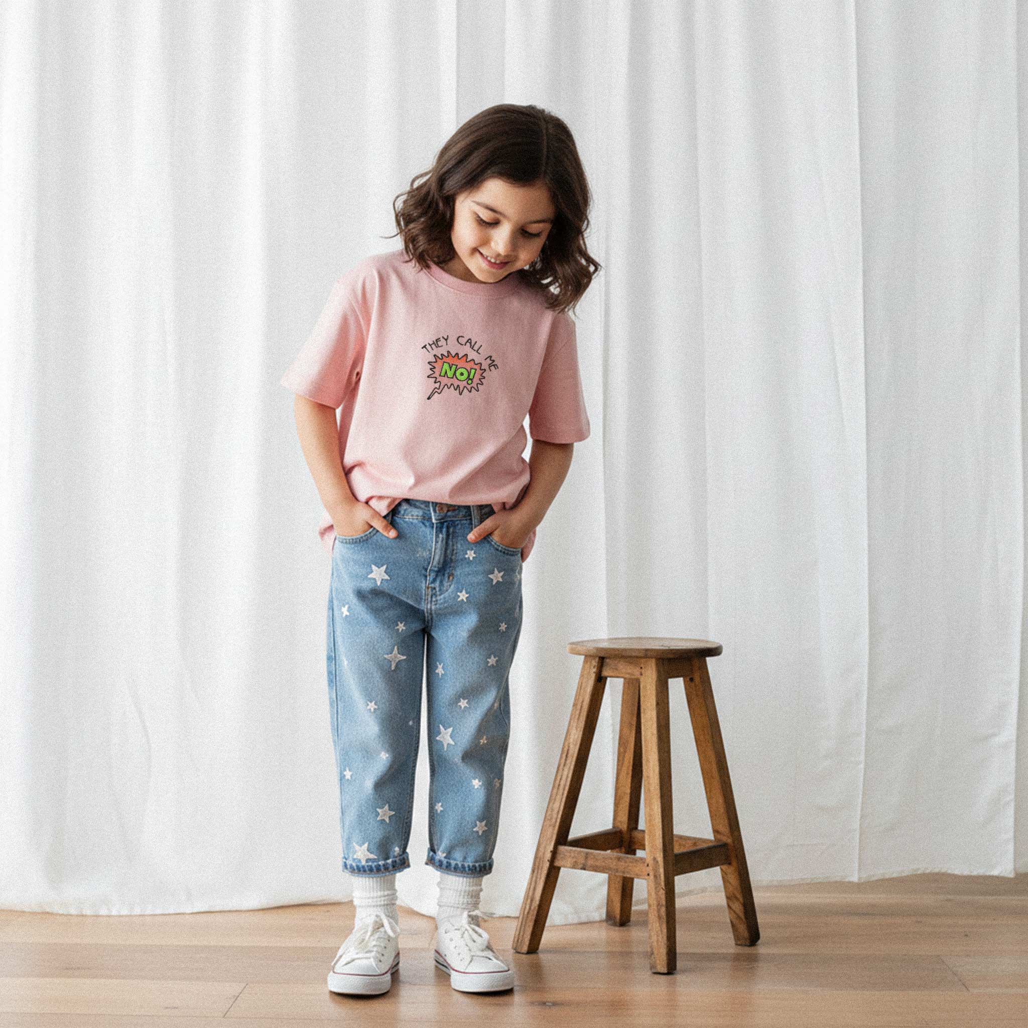 Yıldız desenli jean pantolon ve gül kurusu, 'They Call Me No!' nakışlı oversize tişört giyen kız çocuk, elleri ceplerinde tişörtüne bakarak gülümsüyor.