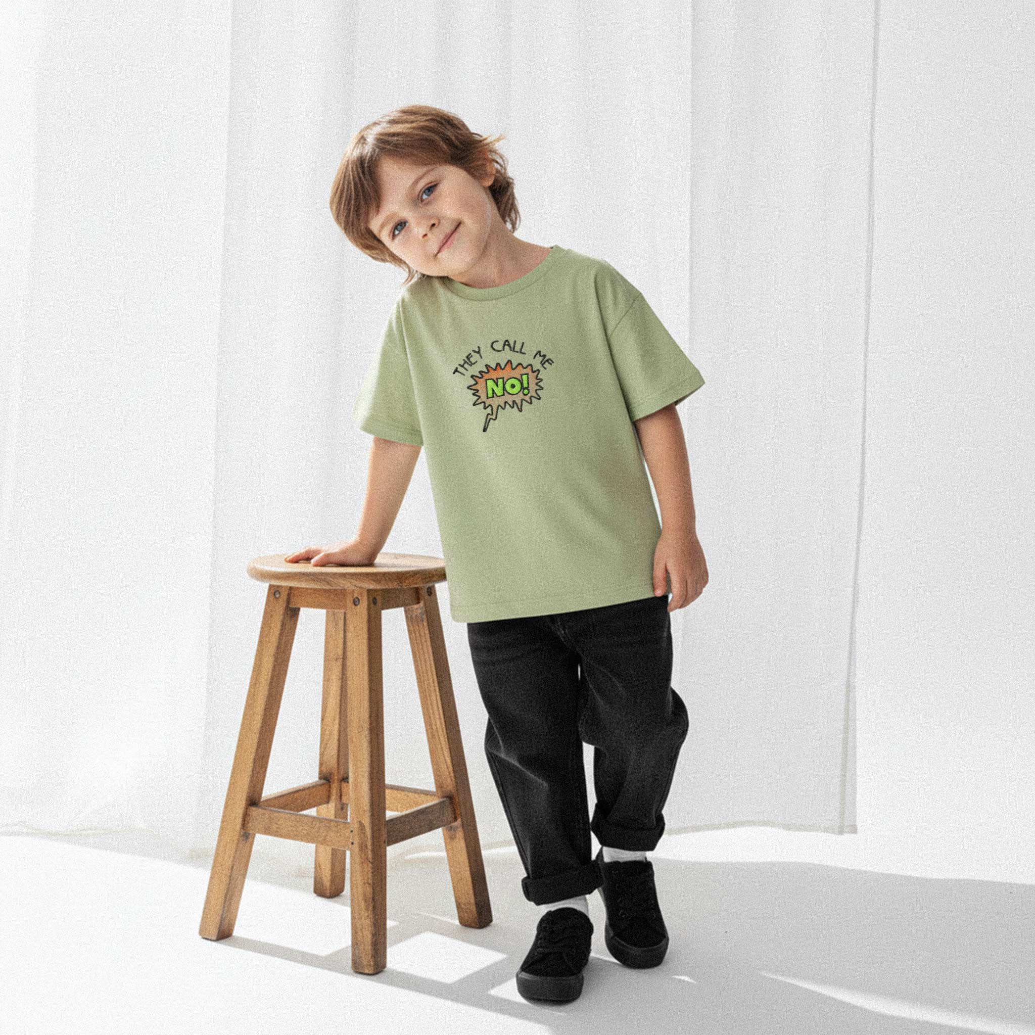 Siyah jean pantolon ve haki yeşili, 'They Call Me No!' nakışlı oversize tişört giyen erkek çocuk, ahşap bir tabureye dayanarak gülümseyerek poz veriyor.