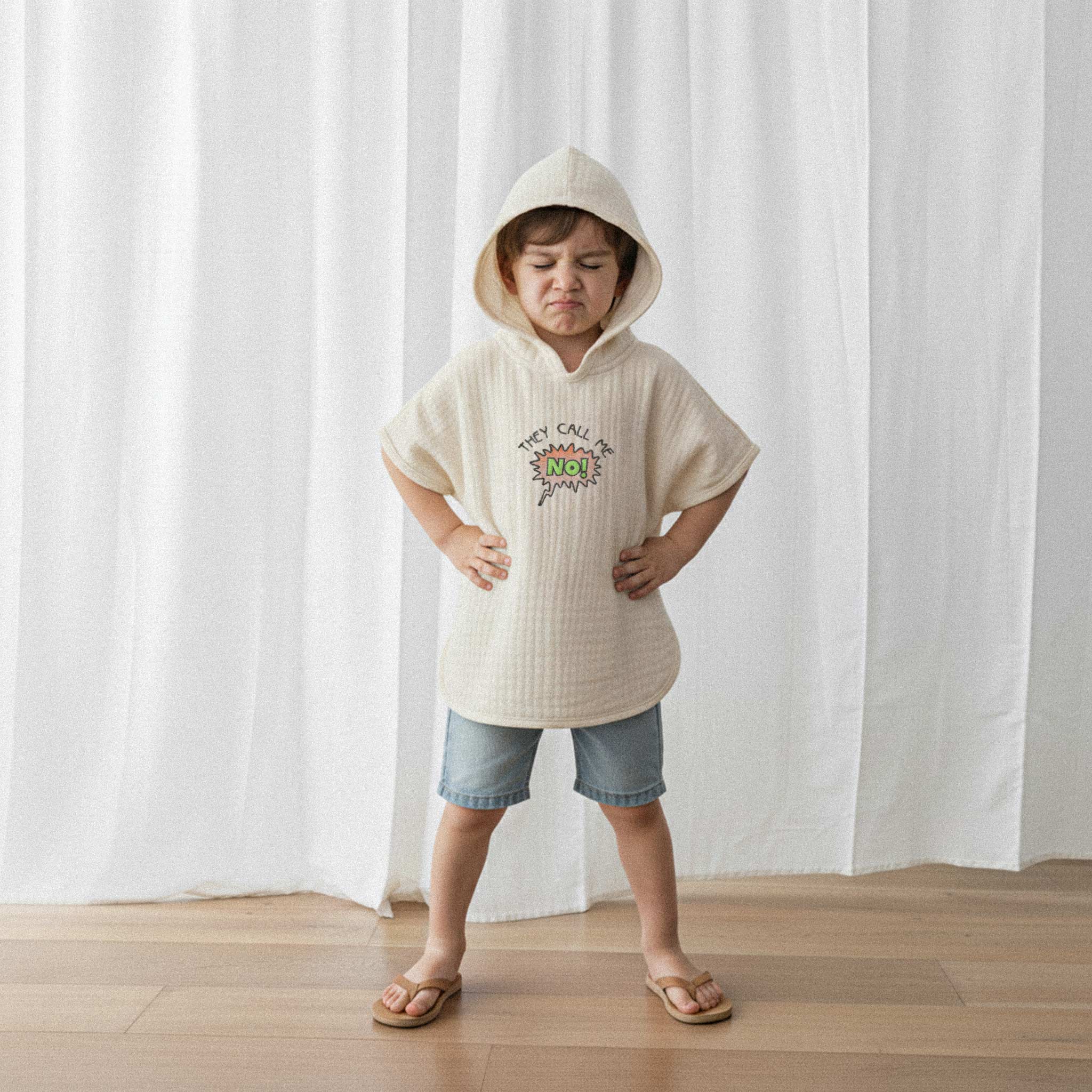 Jean şort ve ekru renkli, 'They Call Me No!' nakışlı kapüşonlu müslin panço giyen küçük erkek çocuk, elleri belinde kızgın bir surat ifadesiyle poz veriyor.