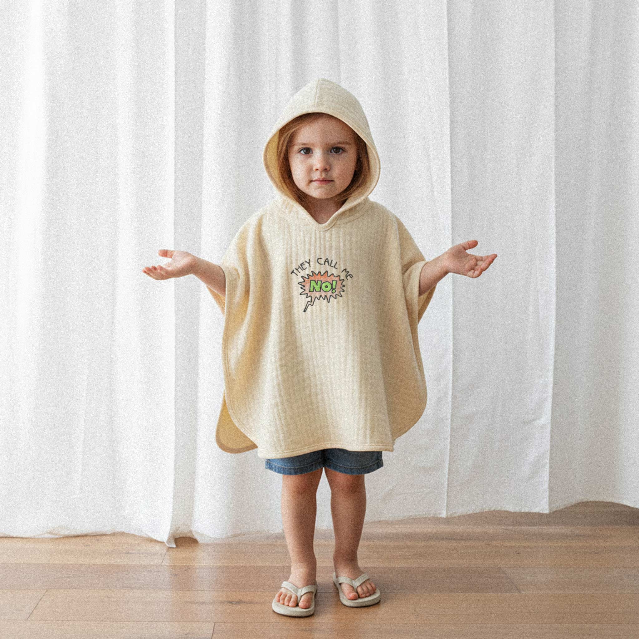 Jean şort ve ekru renkli, 'They Call Me No!' nakışlı kapüşonlu müslin panço giyen küçük kız çocuk, kollarını iki yana açarak sevimli bir poz veriyor.