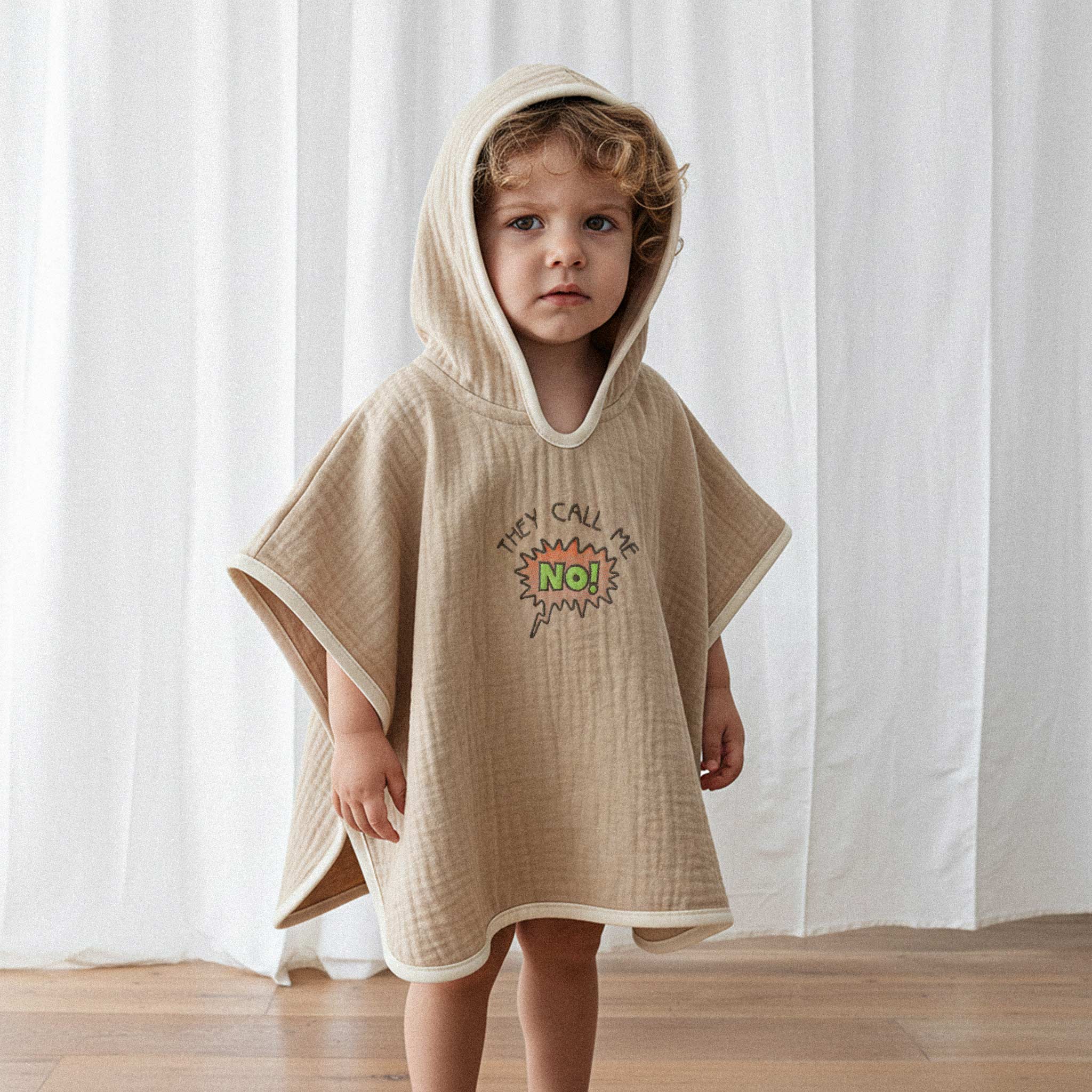Üzerinde somon rengi, 'They Call Me No!' nakışlı kapüşonlu müslin panço bulunan kıvırcık saçlı küçük erkek çocuk, ciddi bir ifadeyle kameraya bakıyor.