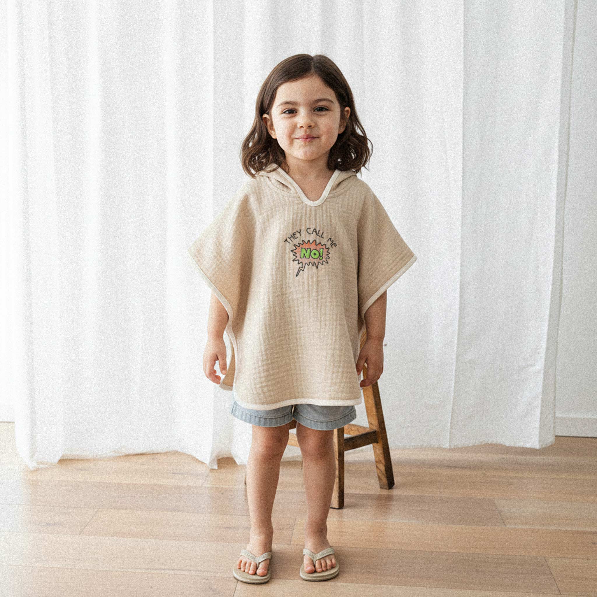 Jean şort ve somon rengi, 'They Call Me No!' nakışlı kapüşonlu müslin panço giyen küçük kız çocuk, gülümseyerek poz veriyor.