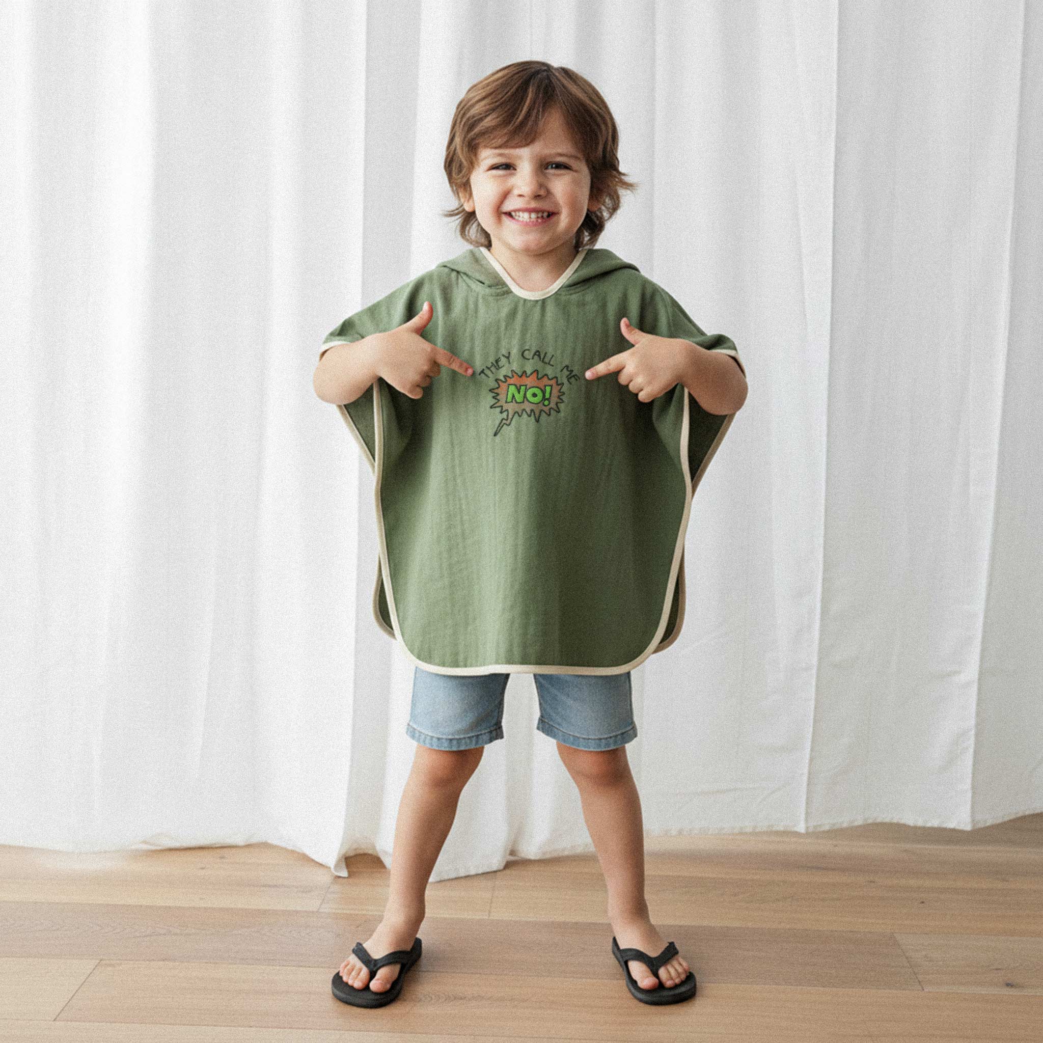Jean şort ve haki yeşili, 'They Call Me No!' nakışlı kapüşonlu müslin panço giyen erkek çocuk, gülümseyerek pançonun üzerindeki yazıyı işaret ediyor.