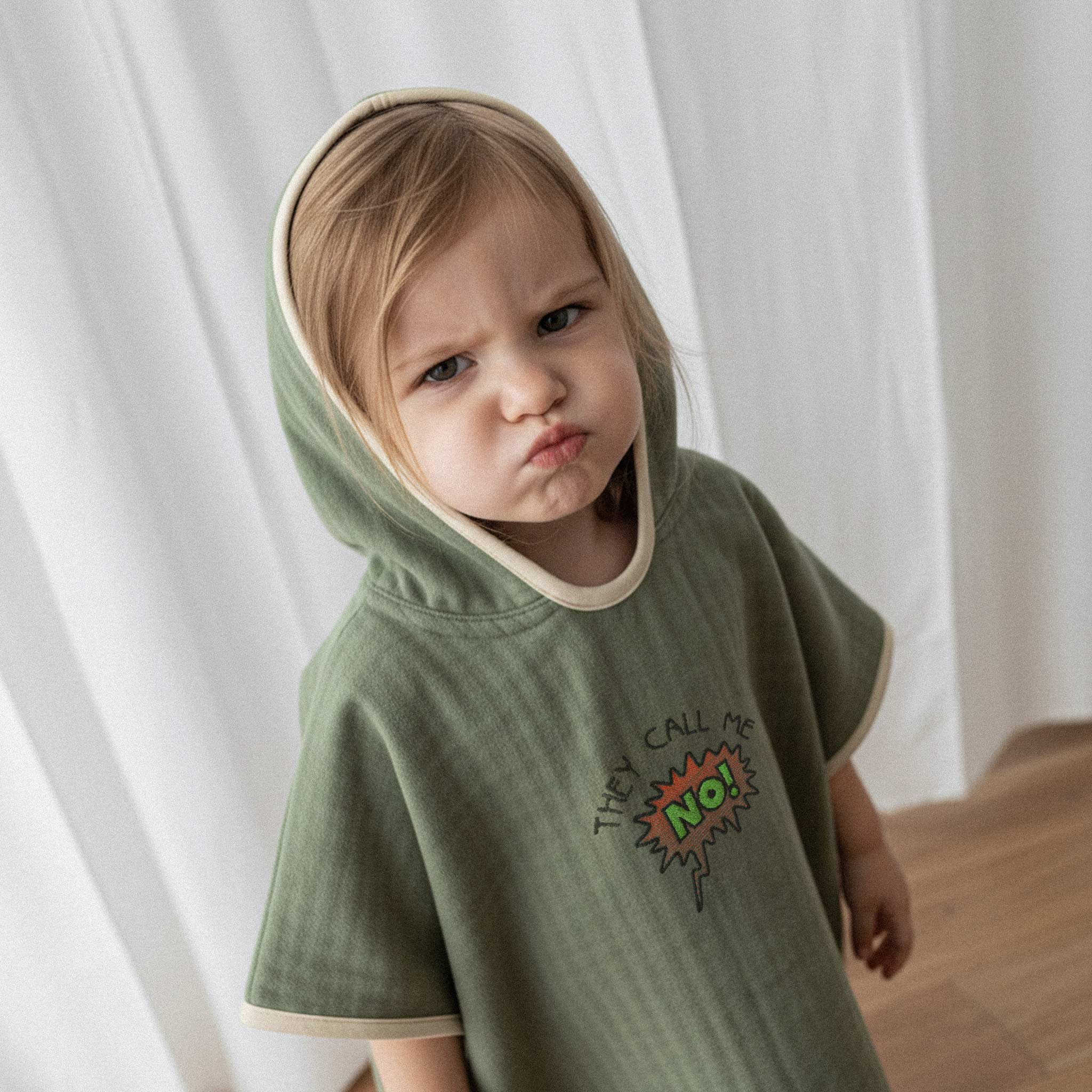 Üzerinde haki yeşili renkli, 'They Call Me No!' nakışlı kapüşonlu müslin panço bulunan küçük kız çocuğun somurtarak baktığı yakın çekim portre.