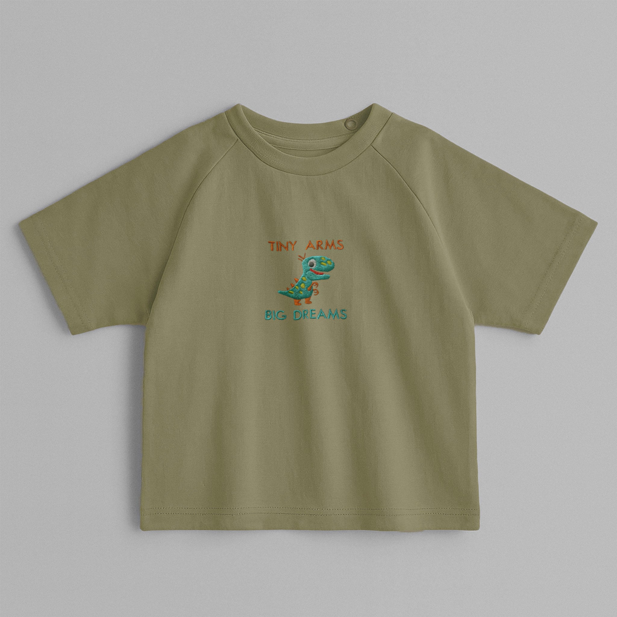 Haki yeşili, omuzdan çıtçıtlı, üzerinde sevimli dinozor nakışı ve 'Tiny Arms Big Dreams' yazısı bulunan pamuklu bebek tişörtü.