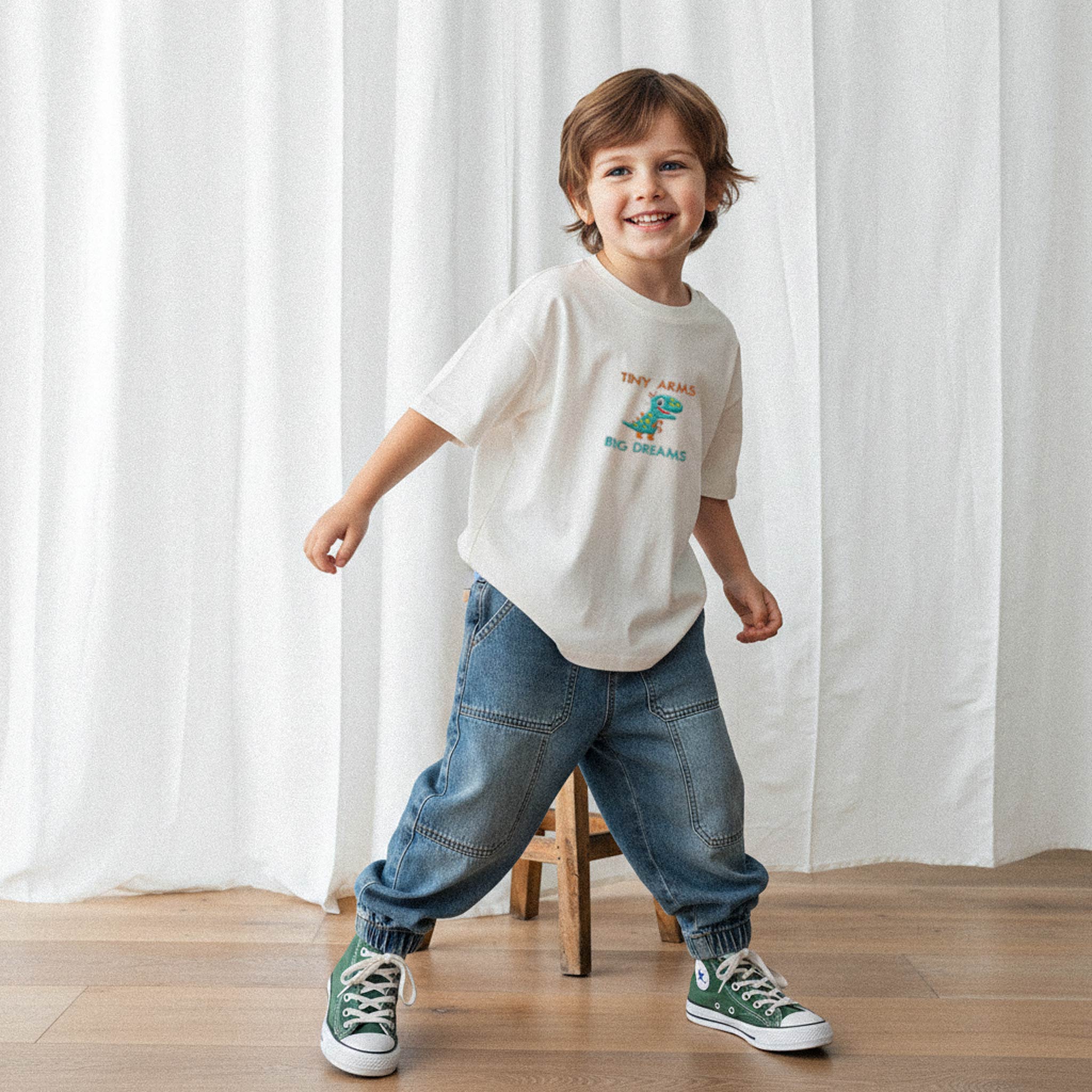 Ekru renkli, dinozor nakışlı oversize tişört ve bol jean pantolon giyen gülen erkek çocuk model.