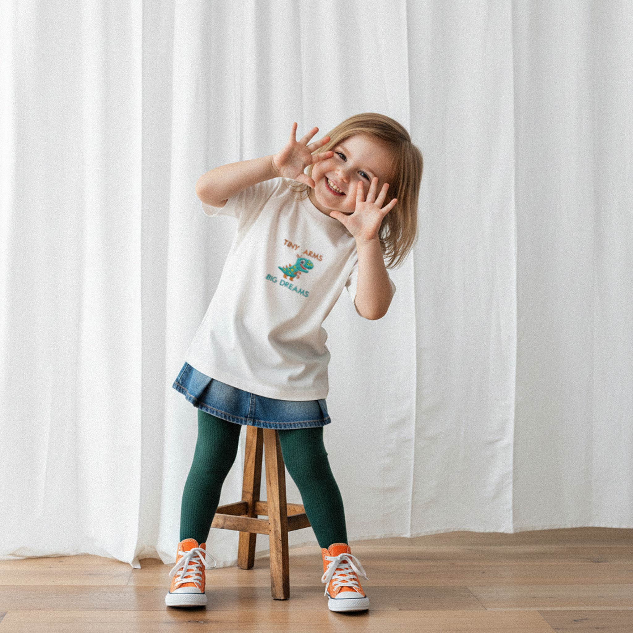 Ekru renkli, dinozor nakışlı tişört giyen, elleriyle yüzünü çerçeveleyerek poz veren neşeli kız çocuk model.