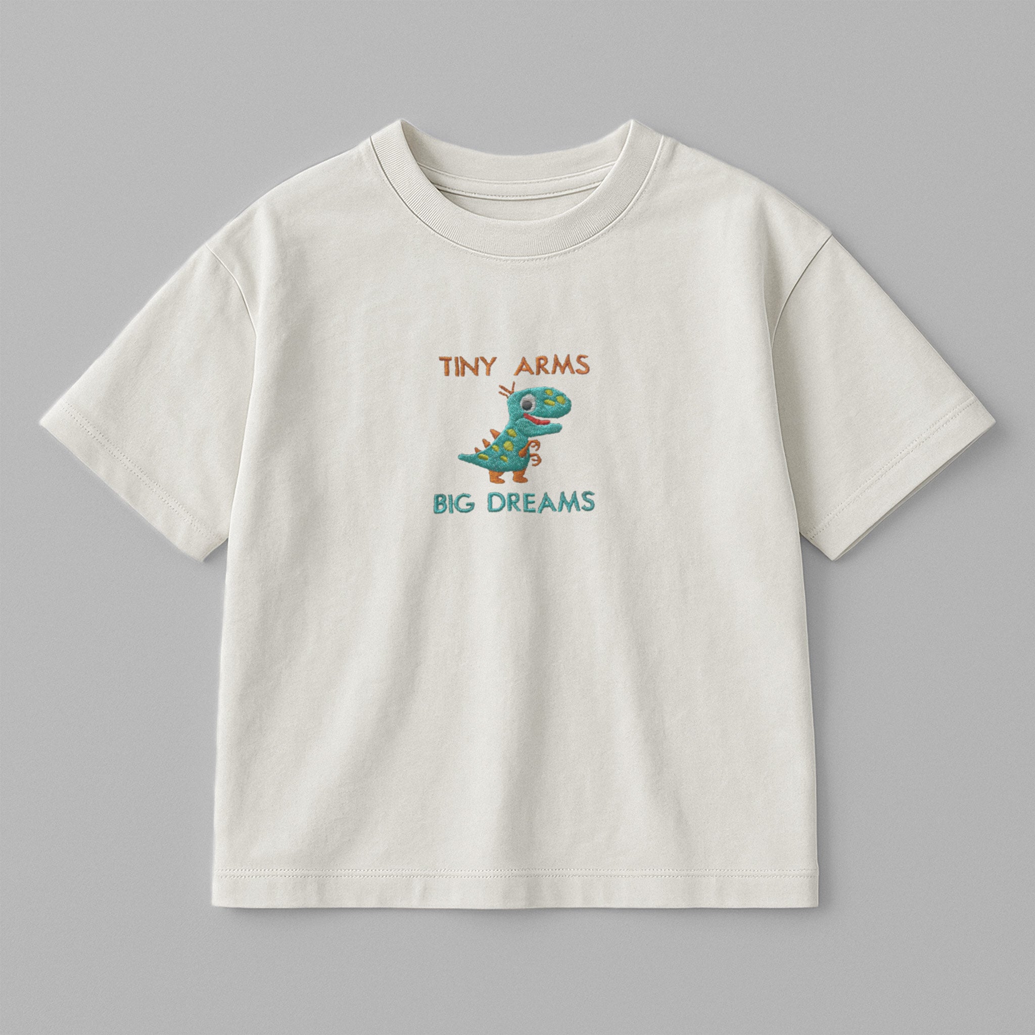 Ekru renkli, oversize kesim, üzerinde sevimli dinozor nakışı ve 'Tiny Arms Big Dreams' yazısı bulunan pamuklu çocuk tişörtü.
