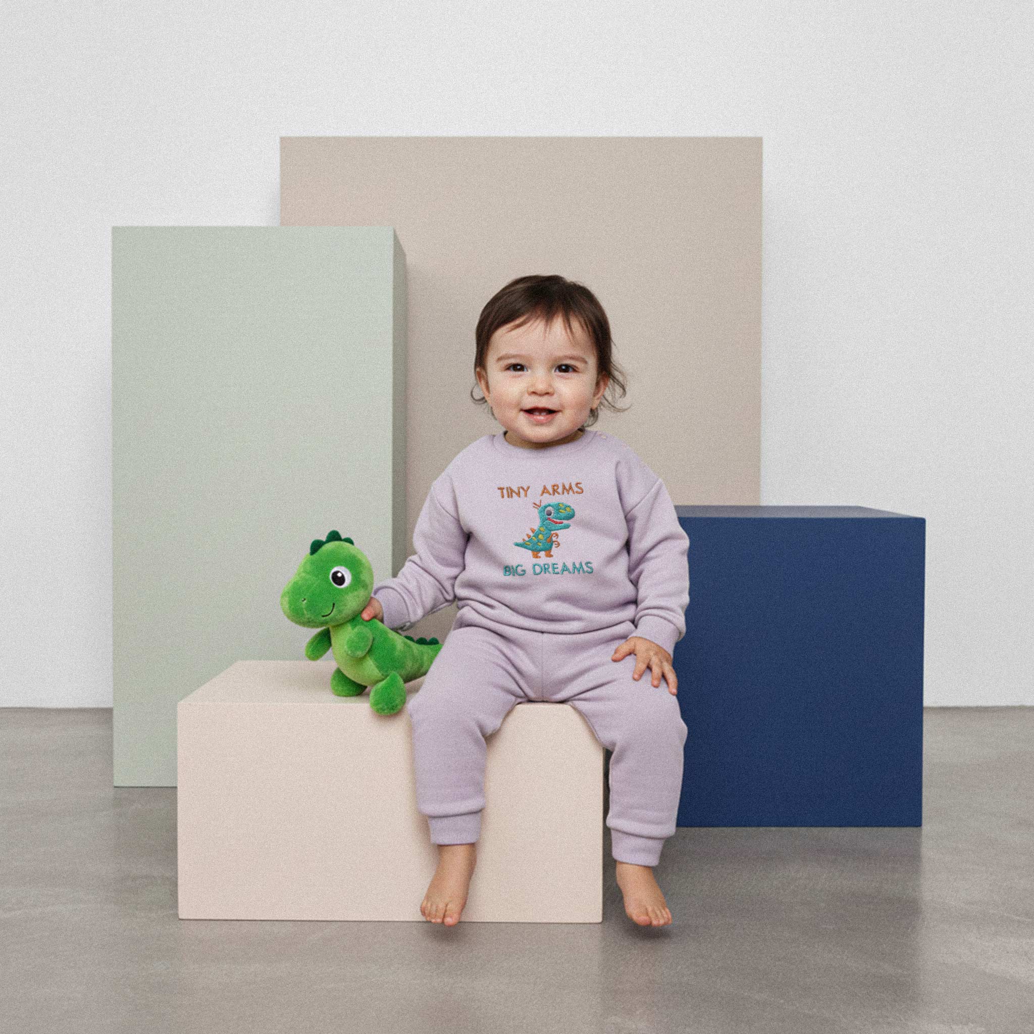 Üzerinde lila renkli, 'Tiny Arms Big Dreams' dinozor nakışlı eşofman takımı bulunan sevimli bebek, elindeki oyuncak dinozorla renkli bir küpün üzerinde oturuyor.