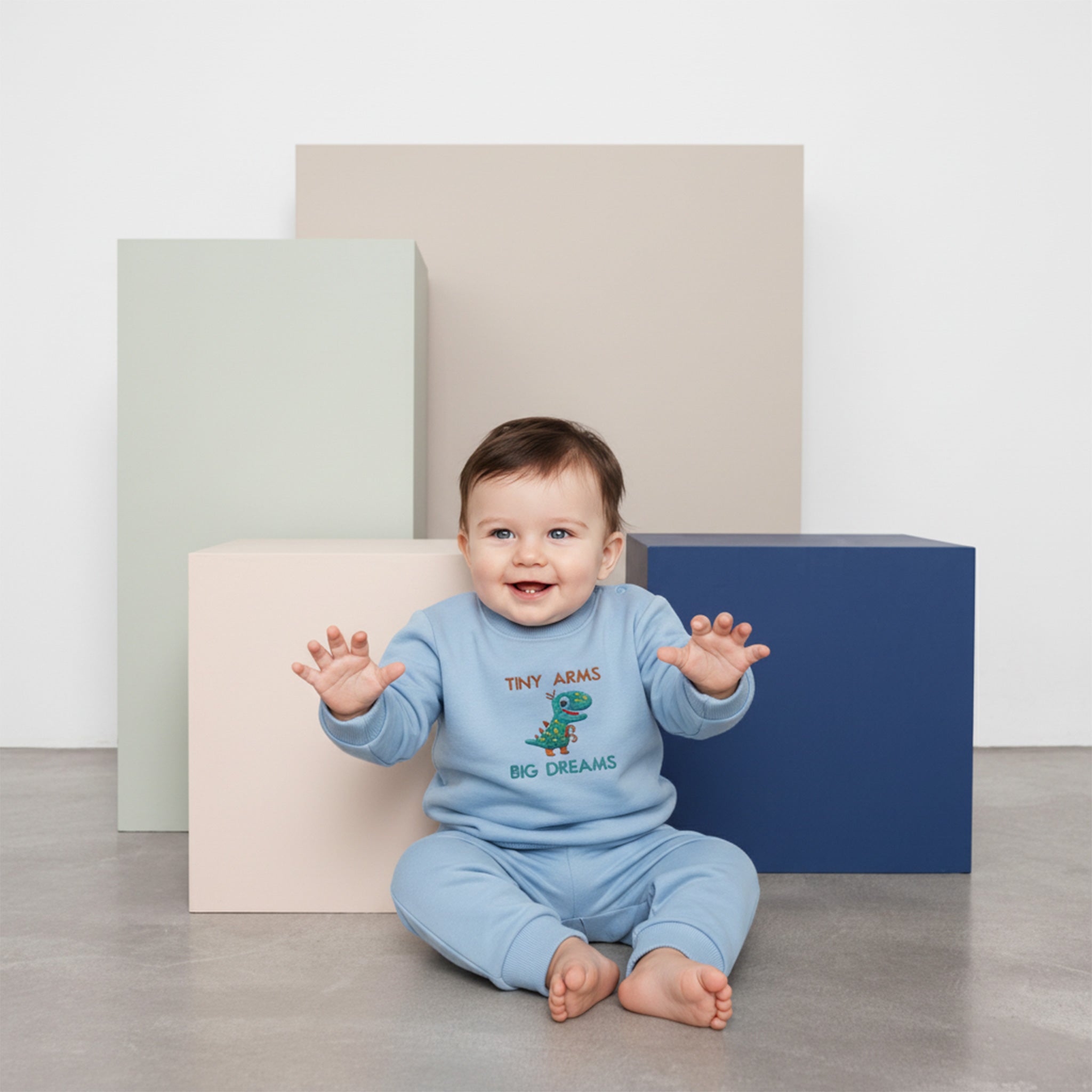 Üzerinde mavi renkli, 'Tiny Arms Big Dreams' dinozor nakışlı eşofman takımı bulunan sevimli bebek, yerde otururken neşeyle ellerini uzatıyor.