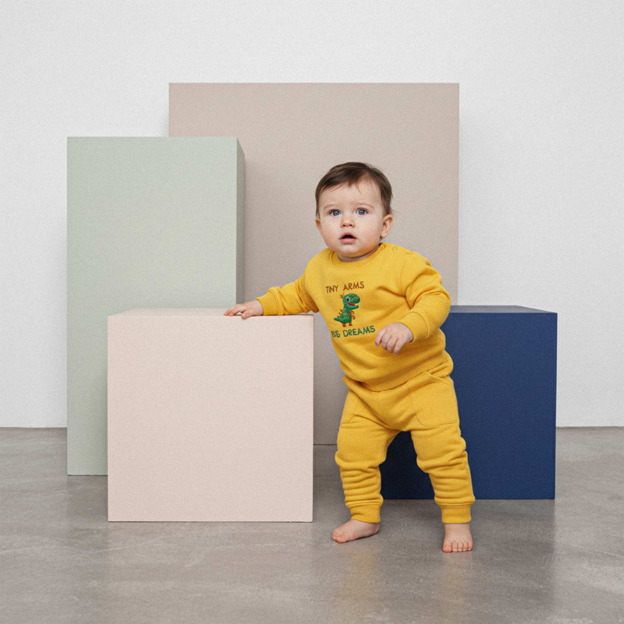 Üzerinde hardal sarısı renkli, 'Tiny Arms Big Dreams' dinozor nakışlı eşofman takımı bulunan sevimli bebek, renkli bir küpe tutunarak ayakta duruyor.