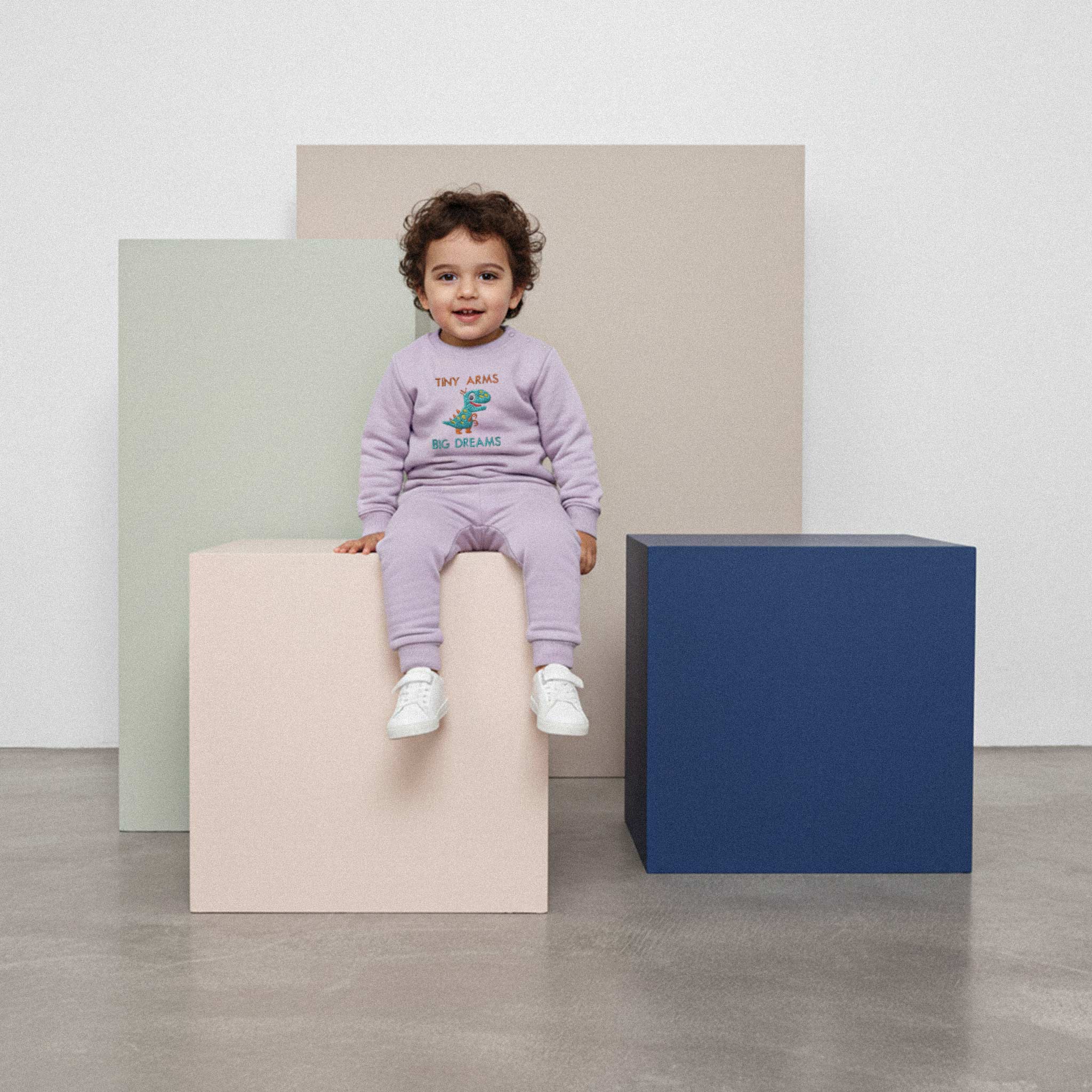 Kıvırcık saçlı, lila renkli 'Tiny Arms Big Dreams' dinozor nakışlı eşofman takımı giyen erkek çocuk, renkli bir küpün üzerinde oturarak gülümsüyor.