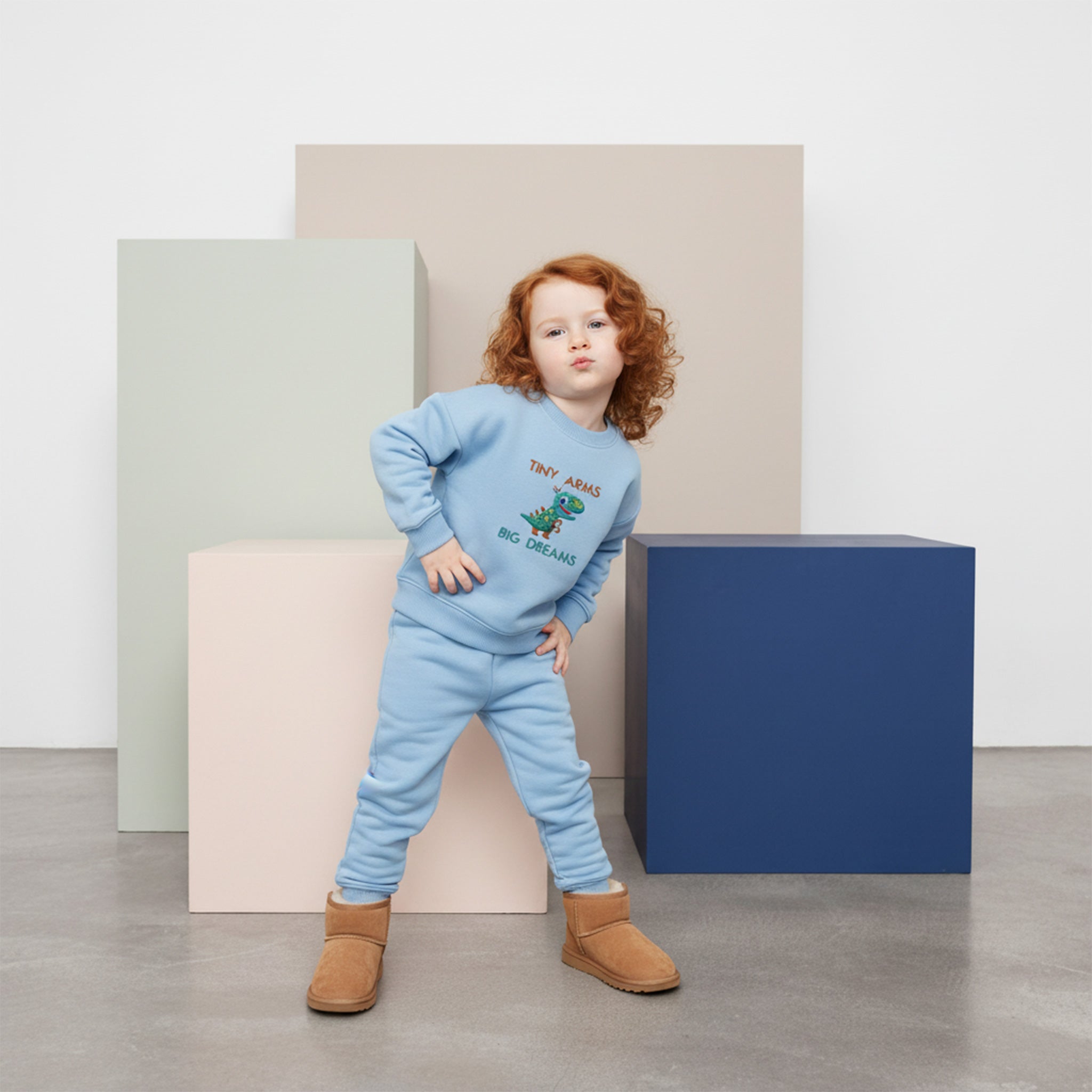 Kızıl saçlı, mavi renkli 'Tiny Arms Big Dreams' dinozor nakışlı eşofman takımı ve botlar giyen çocuk, elleri belinde kendinden emin bir poz veriyor.