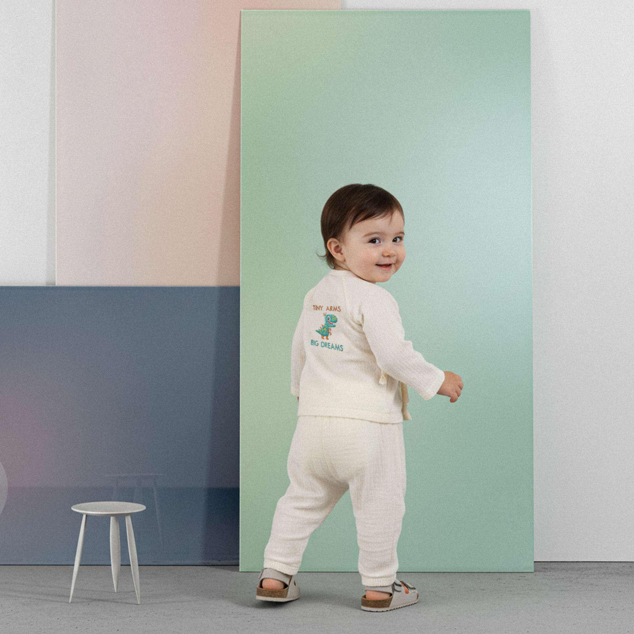 Üzerinde ekru renkli, 'Tiny Arms Big Dreams' dinozor baskılı müslin kimono takımı bulunan sevimli bebek, renkli küplerin önünde ayakta durarak neşeyle gülümsüyor.